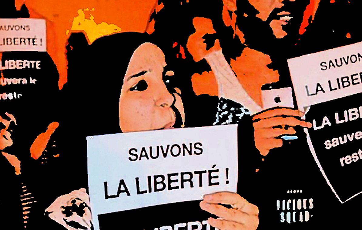 The image depicts a group of people holding up signs with messages in French. The main focus is on two individuals in the foreground. One is wearing a hijab and both are raising their signs high, which read "Sauvons la liberté!" (Save Liberty!). The atmosphere suggests a protest or rally, with a sense of solidarity and determination among the participants. The background is filled with more people and signs, contributing to the overall theme of advocating for freedom.