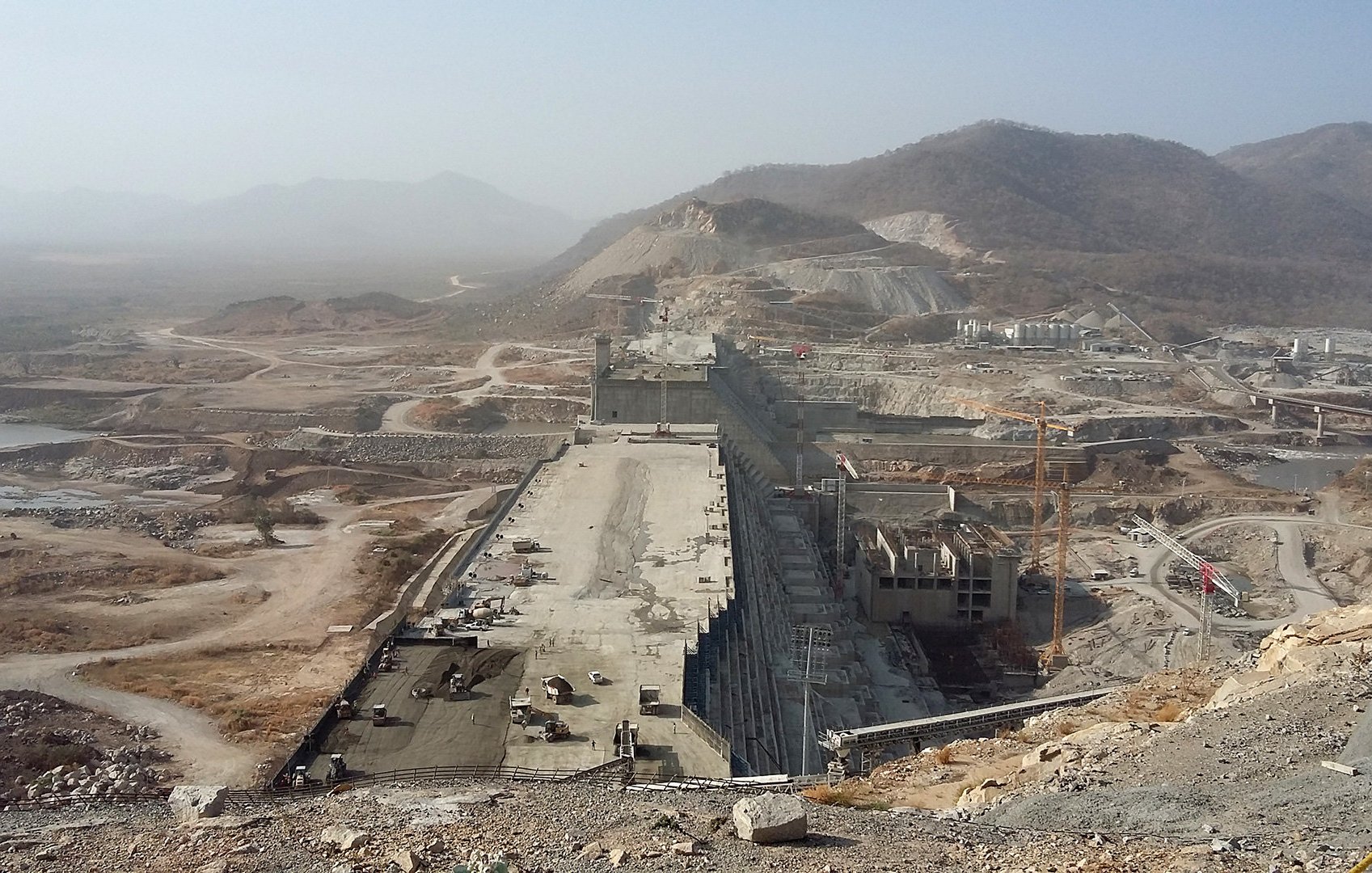The image depicts a large, under-construction dam situated in a mountainous landscape. The dam site features extensive concrete structures and machinery, indicating significant engineering work in progress. Surrounding the site, there are vast areas of earth that have been moved or excavated, along with roads and pathways that suggest ongoing construction activity. The landscape is somewhat arid, with sparse vegetation and mountainous terrain in the background. The visibility is limited, possibly due to haze or dust in the atmosphere.