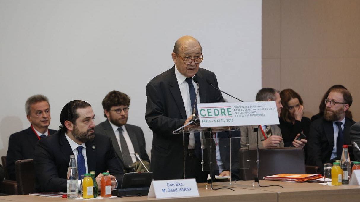 The image depicts a formal conference setting. A middle-aged man, presumably a speaker, is standing at a podium with a sign that reads "CEDRE" and indicates the location and date of the event. He appears to be addressing an audience that includes several men seated behind him, some of whom are engaged in listening or taking notes. The environment has a professional tone, with tables set up and various drinks and papers visible. The atmosphere suggests a significant discussion or presentation, likely focused on economic or policy-related matters.