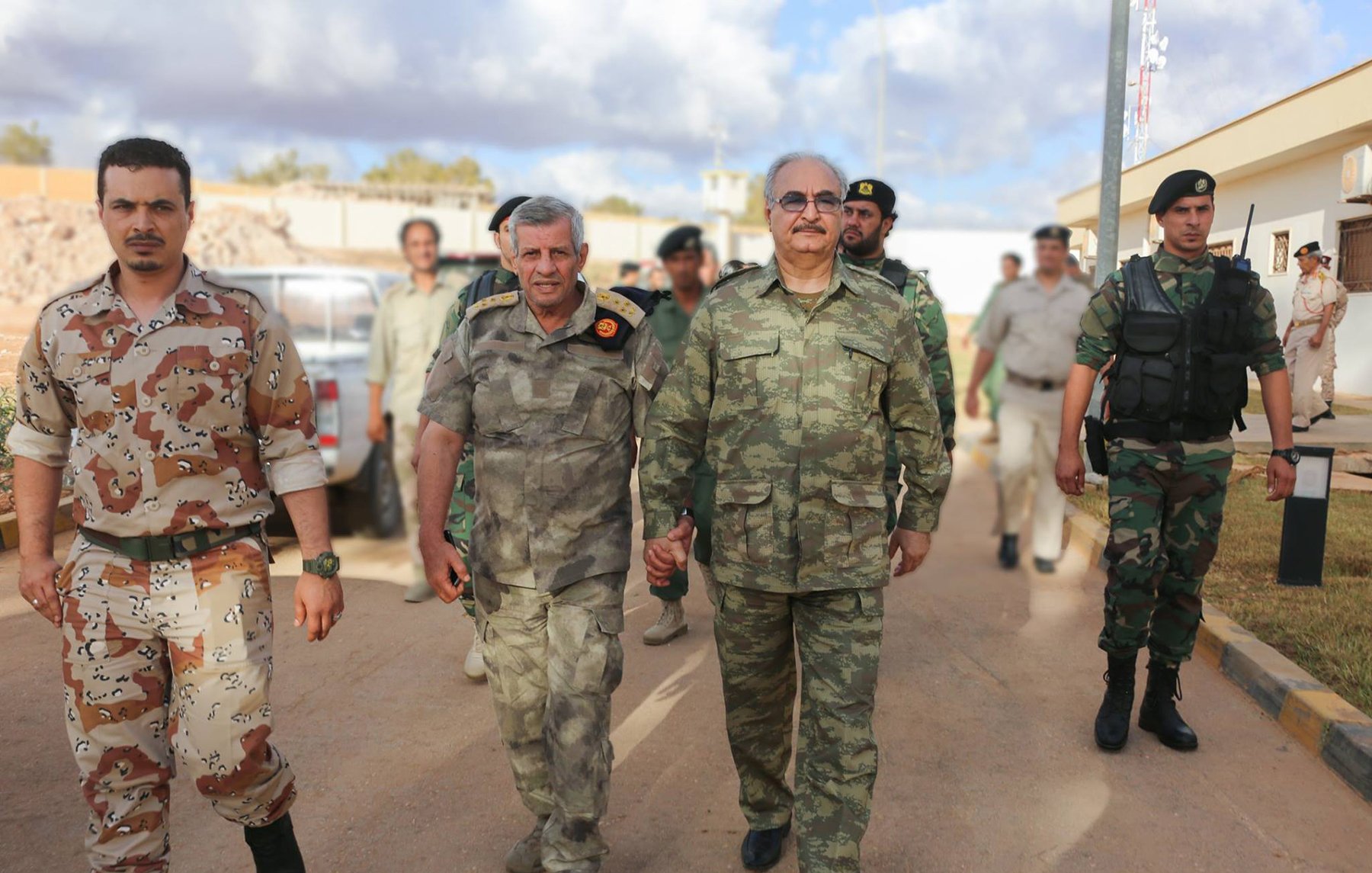 The image depicts a group of military personnel walking together. Two men in the foreground, wearing military uniforms, are holding hands and appear to be engaged in conversation. One is in a camouflage uniform, while the other has a distinctive pattern. They are flanked by soldiers in various military attire in the background. The setting appears to be an outdoor area, possibly a military base, with vehicles and other personnel visible. The sky shows some clouds, indicating daytime.
