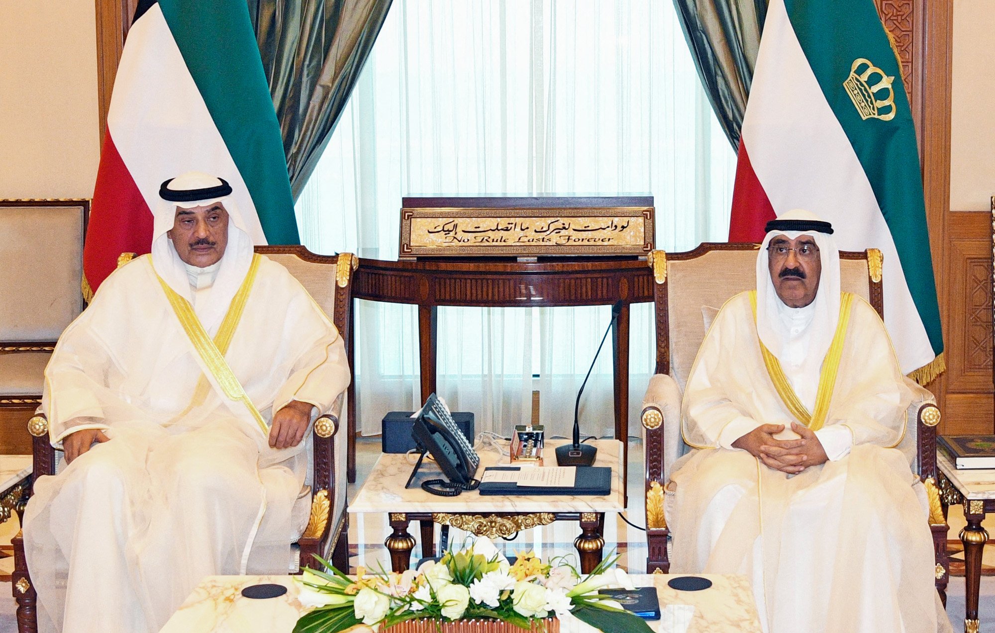The image shows two men seated in a formal setting, likely in a government or diplomatic office. Both are wearing traditional attire, including white thobes and head coverings. They are positioned next to each other, with an ornate decorative background featuring curtains and a rich interior design. On the table in front of them, there are flowers and a few items, including a telephone. Behind them, a plaque with Arabic script can be seen, indicating the formal nature of the meeting.