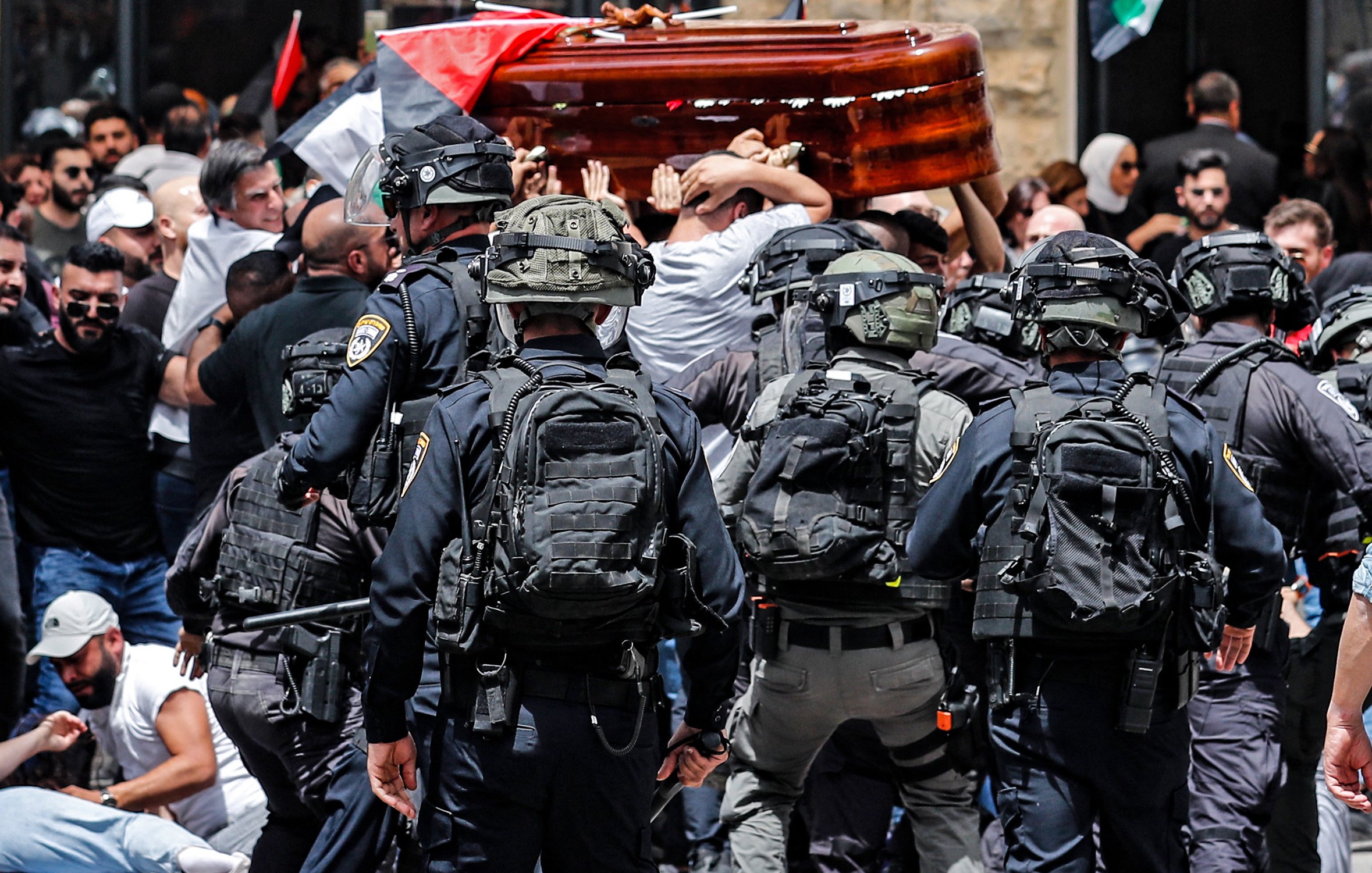 The image depicts a scene of tension and conflict. In the foreground, a group of police officers in tactical gear is facing a larger crowd of people who are mournfully carrying a wooden coffin. The crowd appears to be protesting or commemorating a significant event, with flags visible, including Palestinian flags. The atmosphere is charged, suggesting a clash between the police and the public. Emotions are high as both sides engage in this moment.