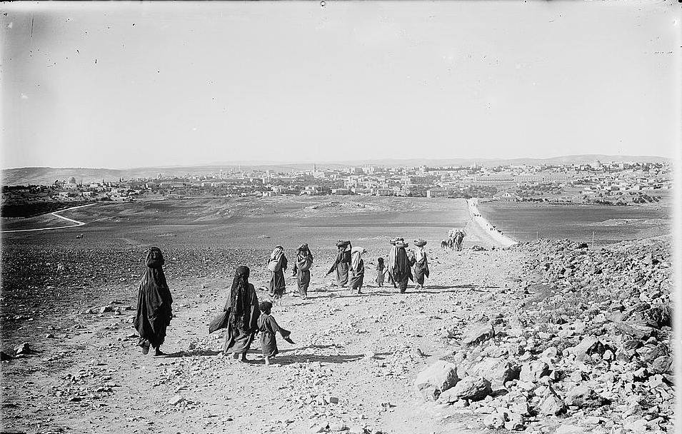 The image depicts a group of people walking along a rocky path in a barren landscape. They appear to be headed toward a distant town or village, which is visible in the background. The group consists of several adults, some of whom are carrying burdens on their heads, and a child walking alongside them. The terrain is dry and rugged, with scattered rocks and sparse vegetation. The sky is clear, and the scene conveys a sense of movement and migration through a rural setting.