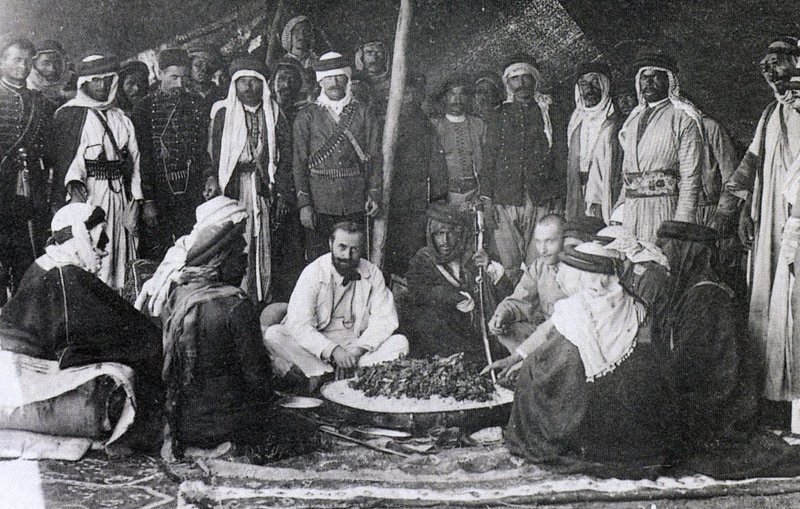 The image depicts a historical gathering of people, likely in a desert or rural setting. In the center, a group of men is seated around a large circular dish, which appears to contain food. They are dressed in traditional attire, with some wearing turbans and others in military or colonial-style uniforms. The background features a tent or canopy, suggesting a communal or ceremonial atmosphere. The mood seems convivial, as the participants engage with one another, possibly sharing a meal or discussing important matters. Overall, the scene reflects cultural practices and social interactions from the past.