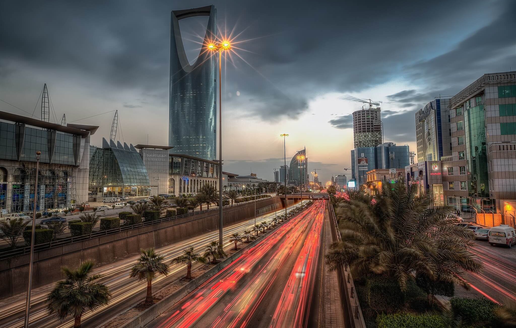 The image depicts a bustling urban scene, likely in a modern city. In the foreground, streaks of light from moving vehicles indicate heavy traffic on a multi-lane road, while palm trees line the sides, adding a touch of greenery. In the background, prominent skyscrapers with reflective glass facades dominate the skyline under a cloudy sky, creating a dramatic atmosphere. The scene captures a blend of modern architecture and vibrant city life, illuminated by streetlights and the fading light of sunset.