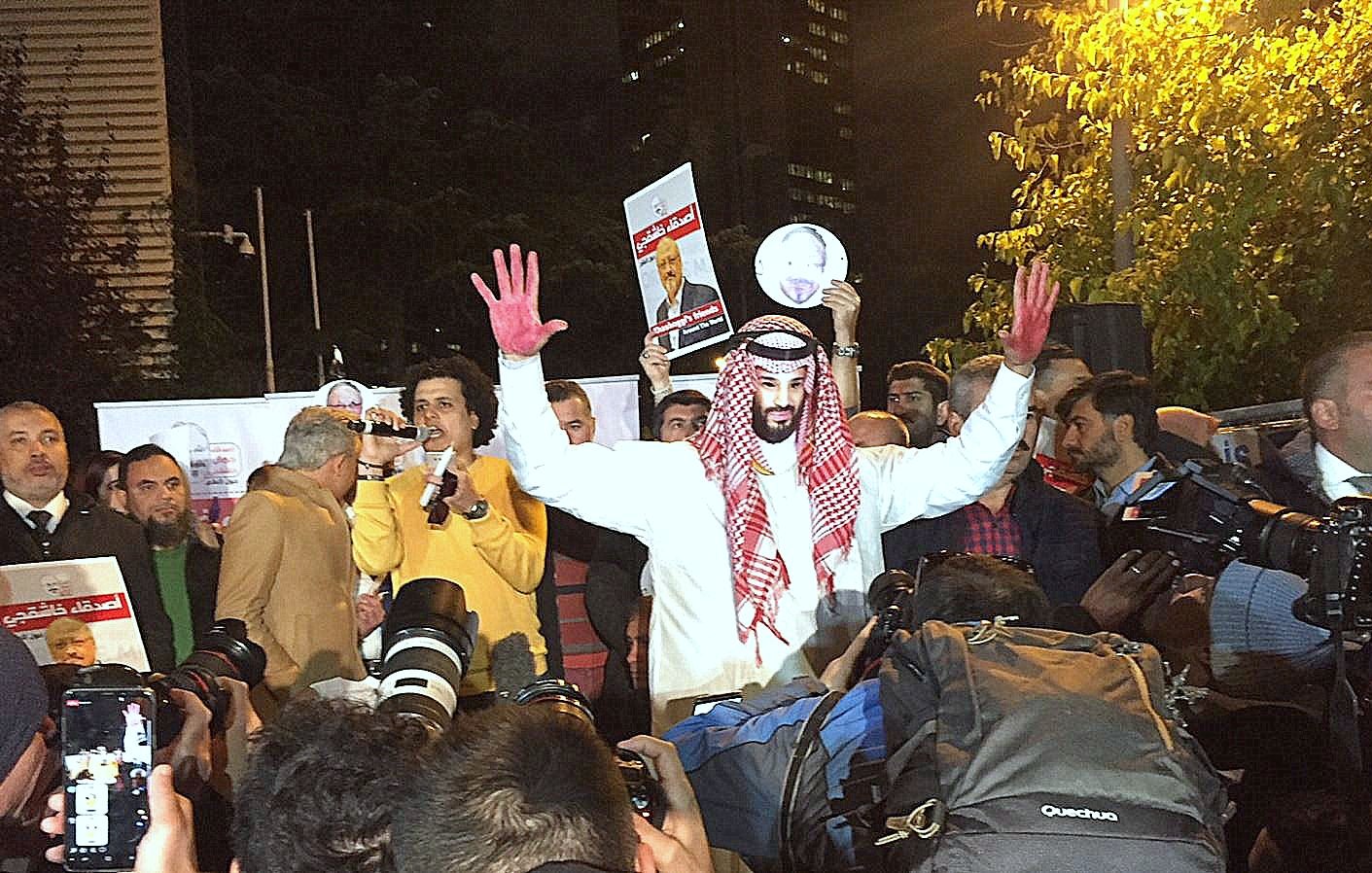 The image shows a group of people gathered for a protest or demonstration. In the foreground, a person wearing traditional attire, possibly a thobe and a headscarf, stands out. They have red paint on their hands, which could symbolize blood or a protest message. The individual is raising their hands and appears to be addressing the crowd or holding up a sign. Surrounding them are numerous cameras and reporters, indicating a media presence. The setting appears to be urban, with buildings visible in the background.
