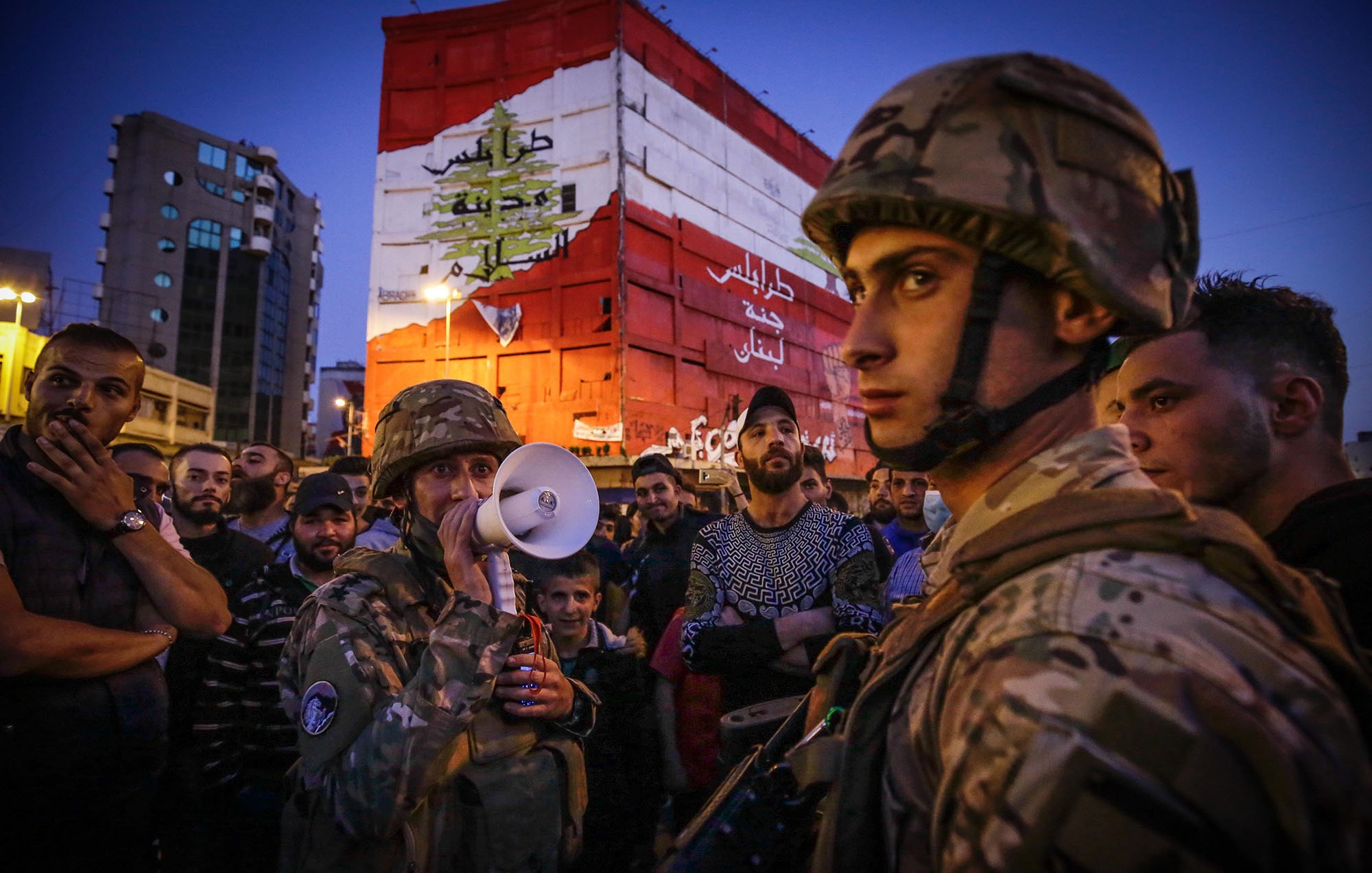 The image depicts a scene of a gathering in an urban environment during the evening. In the foreground, soldiers in military gear, including helmets, are present, with one holding a megaphone, suggesting a communication or announcement to the crowd. The background features a large, colorful wall or mural that displays the Lebanese flag and some Arabic text, likely conveying a political or social message. The crowd surrounding the soldiers appears engaged, with various expressions on their faces, indicating a sense of tension or anticipation in the atmosphere. The setting is illuminated by artificial lighting, hinting at the late hour.