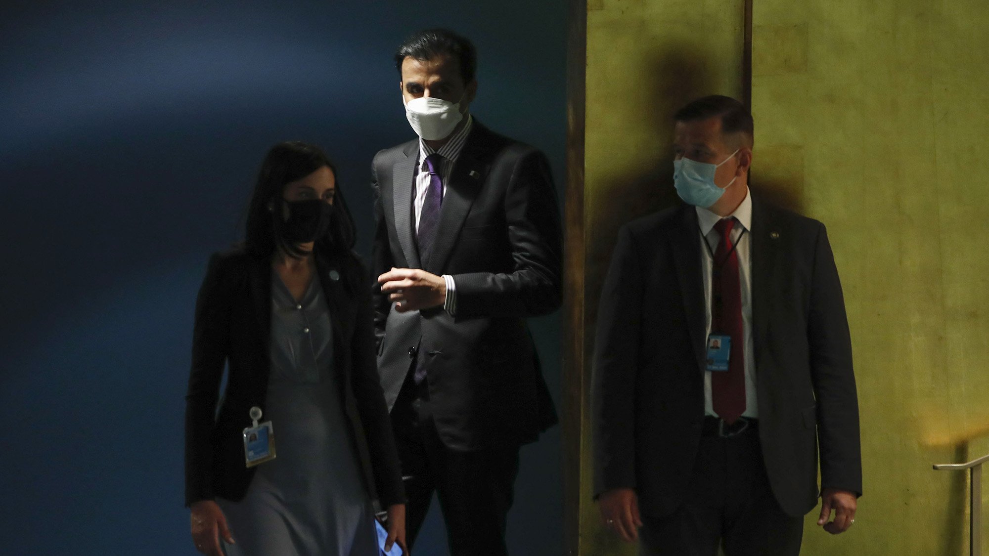 The image shows three individuals in a setting that appears to be a formal environment, likely related to an official event or meeting. Two of them are wearing masks. One person is dressed in a suit and appears to be looking at something on their phone. The other two individuals are also formally dressed, with one wearing a blazer and the other in a suit and tie. They seem to be in a hallway or near a door, suggesting they are entering or exiting a space. The overall atmosphere conveys a sense of professionalism and seriousness.