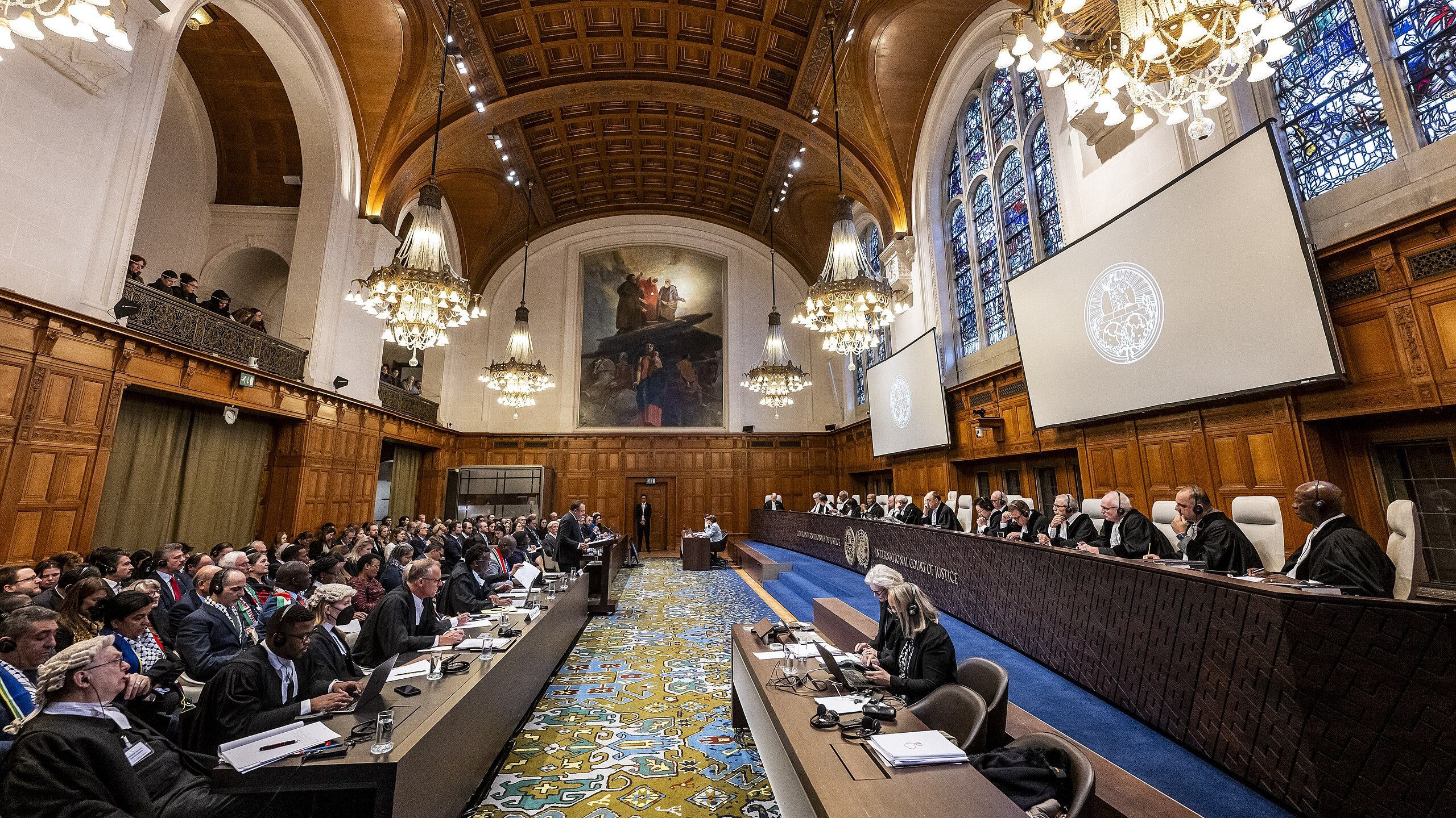 The image depicts an interior of a grand courtroom, likely in an international court such as the International Court of Justice. The room features high wooden ceilings with intricate designs and ornate chandeliers providing warm lighting. Large windows with stained glass allow natural light to filter in. In the foreground, there are rows of judges seated on a raised platform, alongside legal representatives and observers. The layout includes a long table with officials on one side and a broad audience on the other. The design of the room is both formal and historic, emphasizing the importance of the proceedings taking place.
