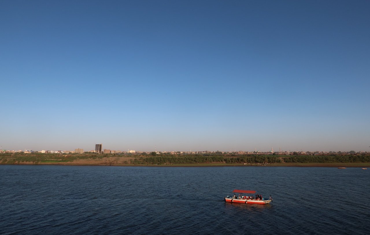 The image depicts a serene river landscape under a clear blue sky. In the foreground, a small boat with a red canopy is floating on the water, which reflects the sky. In the background, there is a sandy shore lined with greenery, and a distant cityscape can be seen, with buildings rising against the horizon. The overall scene conveys a peaceful and picturesque atmosphere.