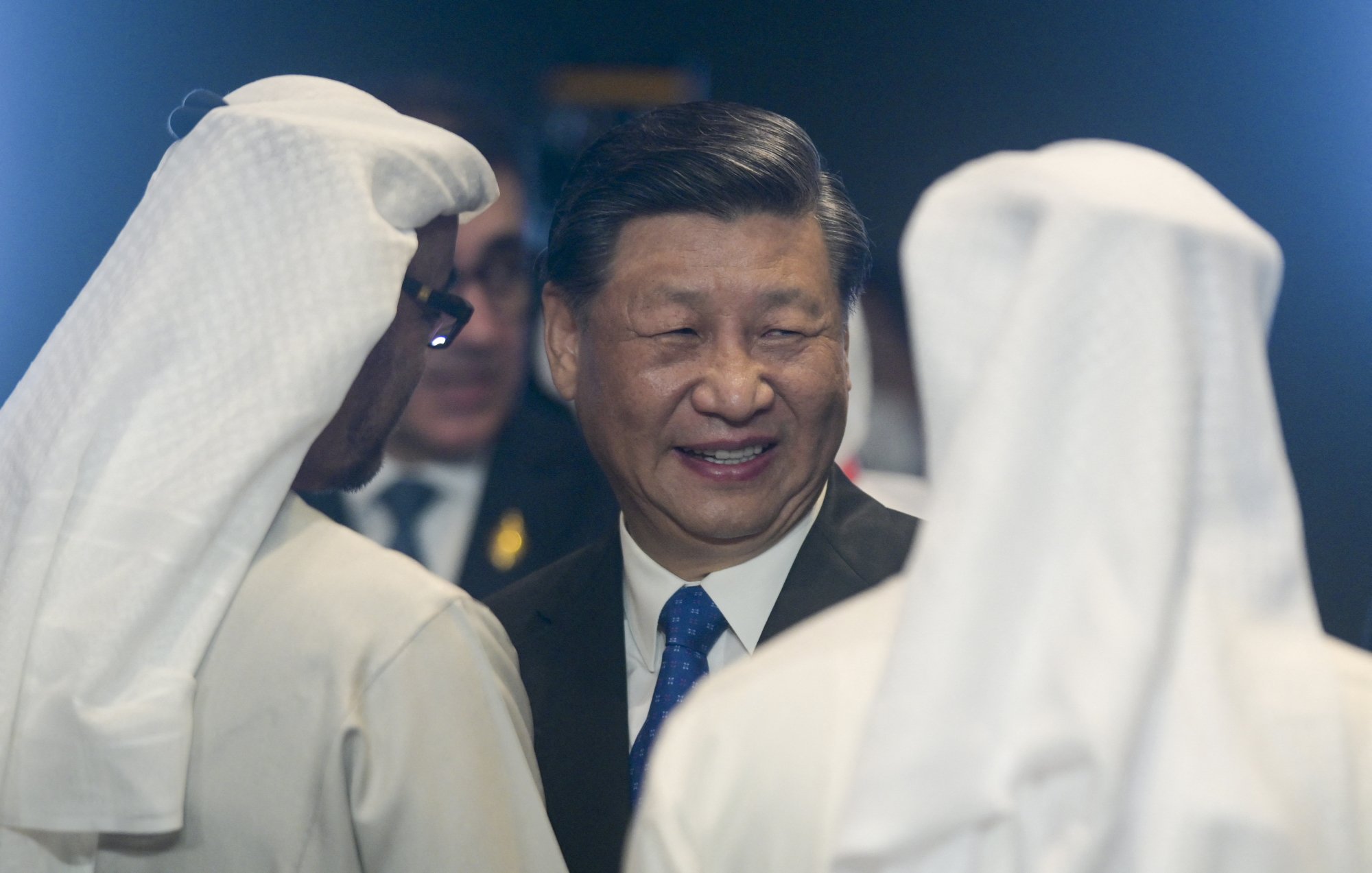 The image depicts a smiling man in a suit engaging in conversation with two individuals wearing traditional white Middle Eastern attire, including headscarves. The setting feels formal and diplomatic.