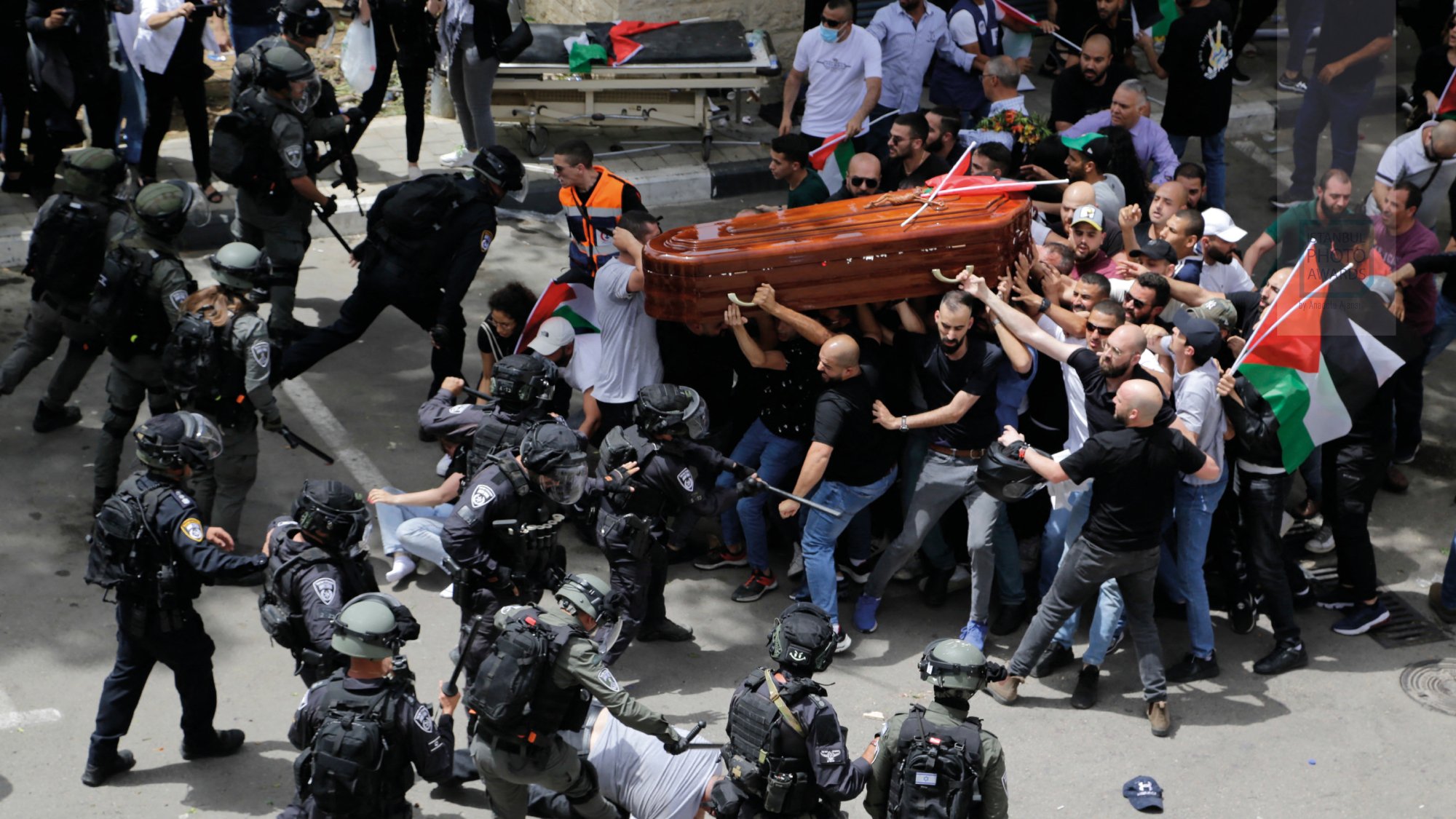 A crowd confronts police while carrying a coffin; flags and tension are visible.