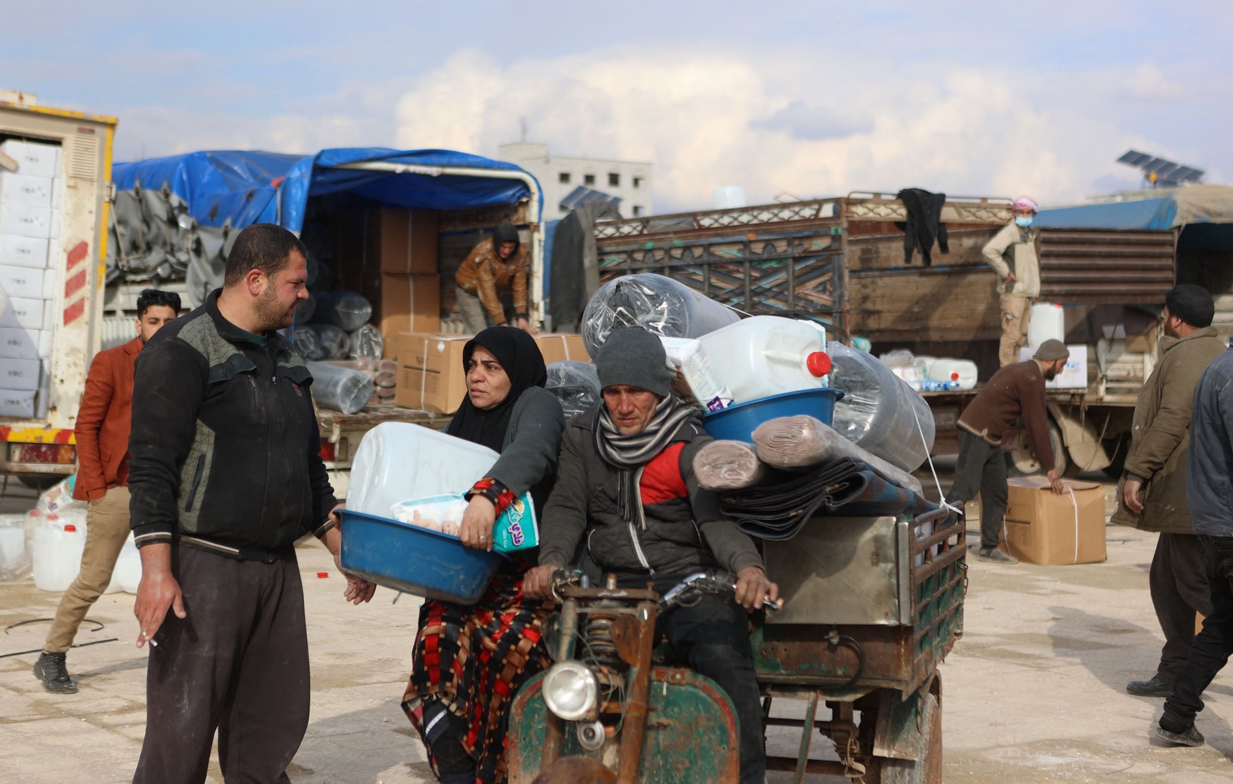 The image shows a busy scene where people are engaged in various activities, likely in a market or a distribution area. In the foreground, a man and a woman are interacting next to a small vehicle loaded with large containers, possibly for water or other supplies. The woman is dressed in traditional clothing and appears to be holding a tray, while the man seems to be dressed in casual attire. In the background, several people are seen unloading boxes and organizing goods, with a truck and some tents visible. The setting has a somewhat informal atmosphere, suggestive of a community effort related to aid or distribution.