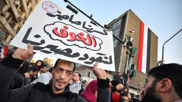 The image depicts a crowd gathered in an urban setting, likely during a protest or demonstration. In the foreground, a man holds a large sign with Arabic text, which translates to "Our demands are not to be ignored." In the background, there is a building displaying the Egyptian flag, suggesting a context related to political or social issues in Egypt. The scene conveys a sense of activism and public expression, with people rallying for their rights or changes in governance.