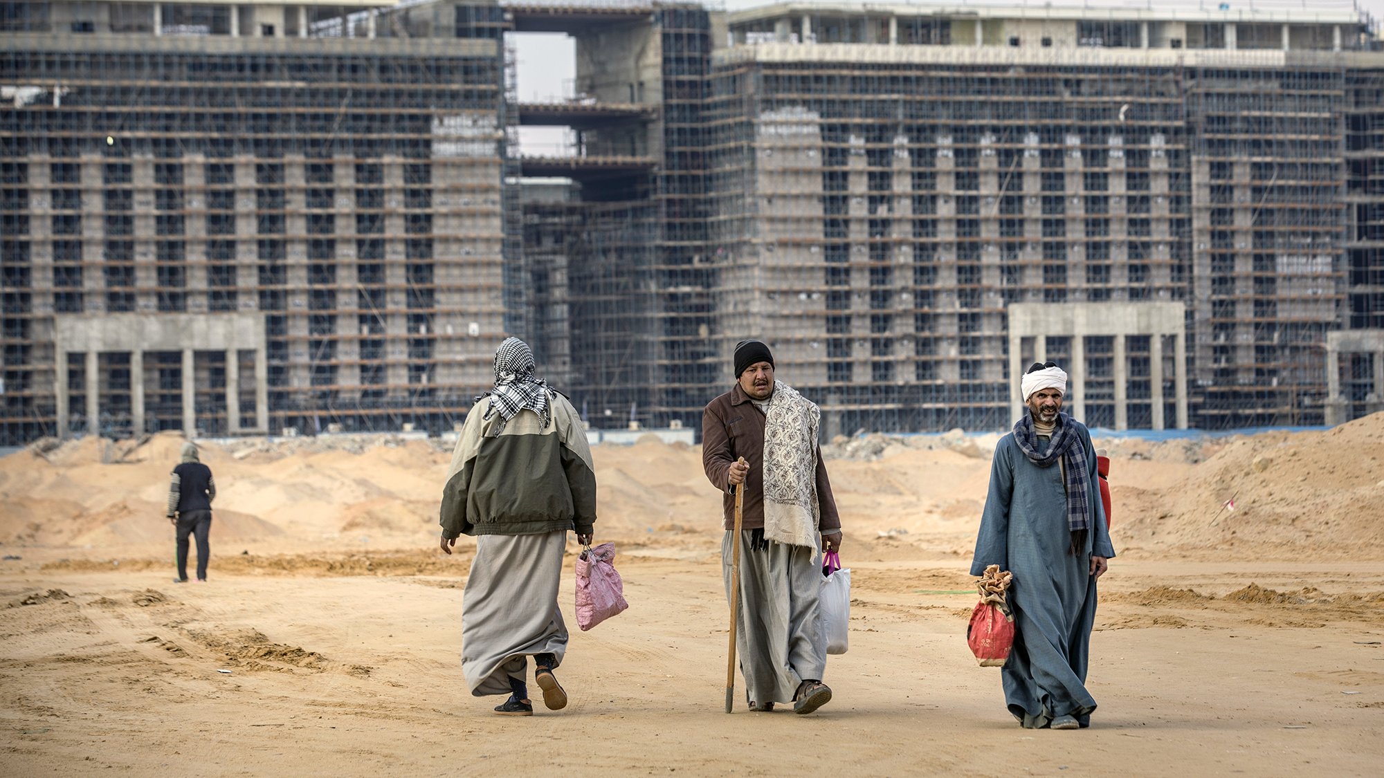 The image depicts a construction site with large, partially completed buildings in the background. In the foreground, four individuals are seen walking on a sandy terrain. They are dressed in traditional attire, including long robes and head coverings. Some are carrying bags, and one person is using a cane. The overall scene reflects a blend of activity and the unfinished nature of the construction, suggesting a developing urban area.