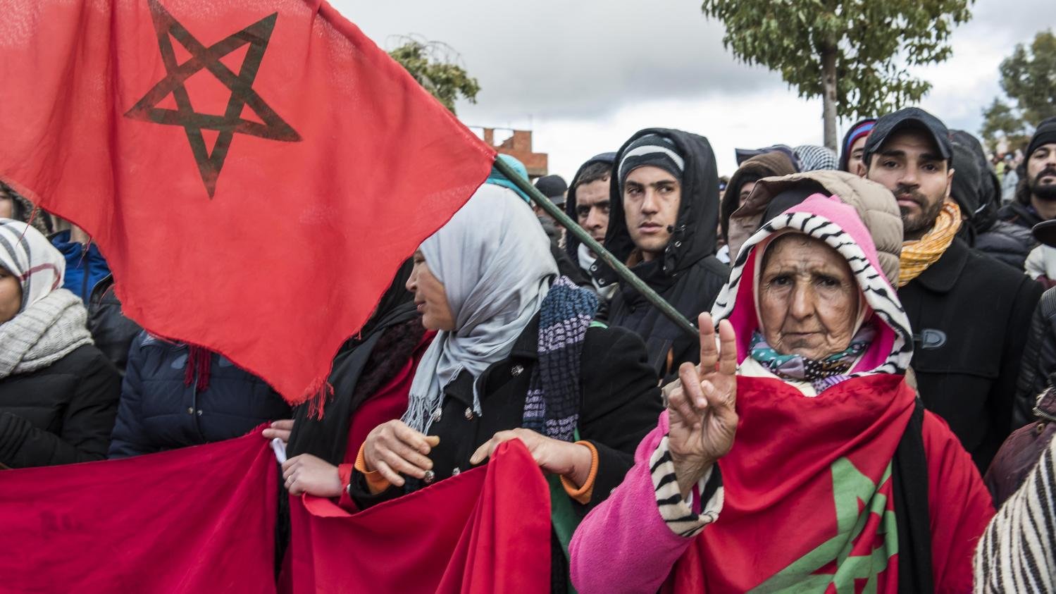 The image depicts a group of people participating in a demonstration. In the foreground, an older woman holds a flag featuring the Moroccan national emblem and makes a peace sign with her hand. She is surrounded by others who are also holding flags and banners, all dressed for colder weather. The atmosphere suggests a sense of solidarity among the demonstrators, with the crowd positioned against a backdrop of cloudy skies.
