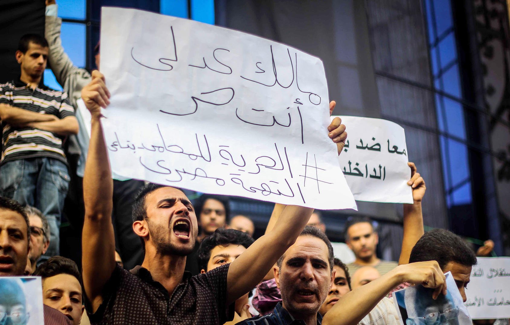 The image depicts a crowd of protestors holding signs during a demonstration. One individual is prominently featured, passionately expressing their views. The sign reads a message in Arabic, which translates to a call for freedom and justice. The crowd appears engaged and determined, highlighting the intense atmosphere of the protest. Various faces in the background show a mix of emotions, reflecting the seriousness of the situation.
