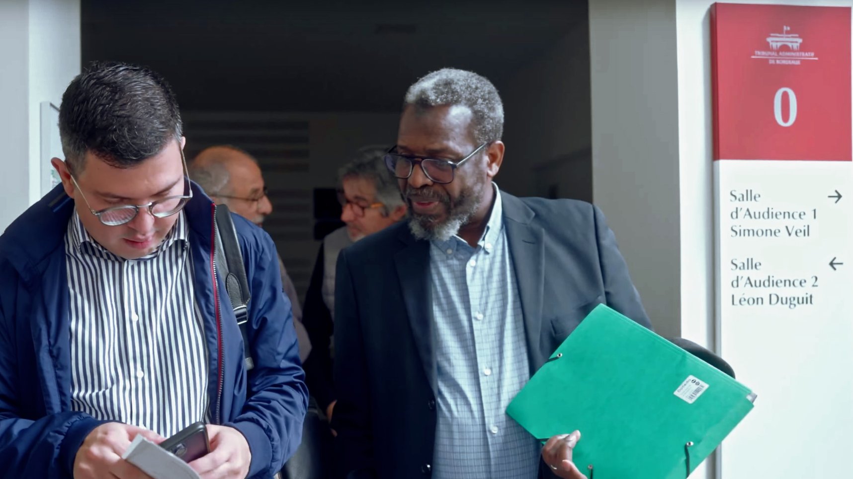 The image shows two men engaged in conversation as they exit a building. One man, wearing glasses and a navy jacket, is looking at his phone while the other, dressed in a black suit, is holding a green folder. In the background, there are other people visible, indicating a busy environment, possibly a court or conference setting. The signage indicates different audience rooms, adding context to the location.