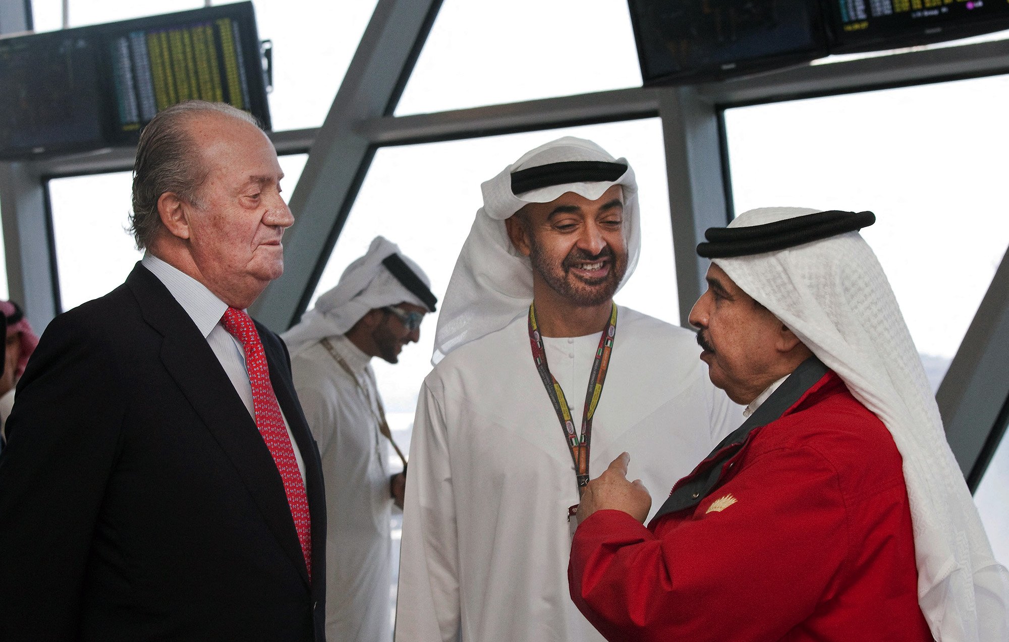 The image shows a gathering of three men engaged in conversation. Two of them are dressed in traditional attire, typical of Middle Eastern cultures, with one wearing a white kandura and the other in a black and white headdress. The third man is in formal Western attire with a suit and a red tie. The background suggests an airport or a similar setting, indicated by the large windows and digital displays. The expressions of the men appear to be friendly and engaged in a dialogue.