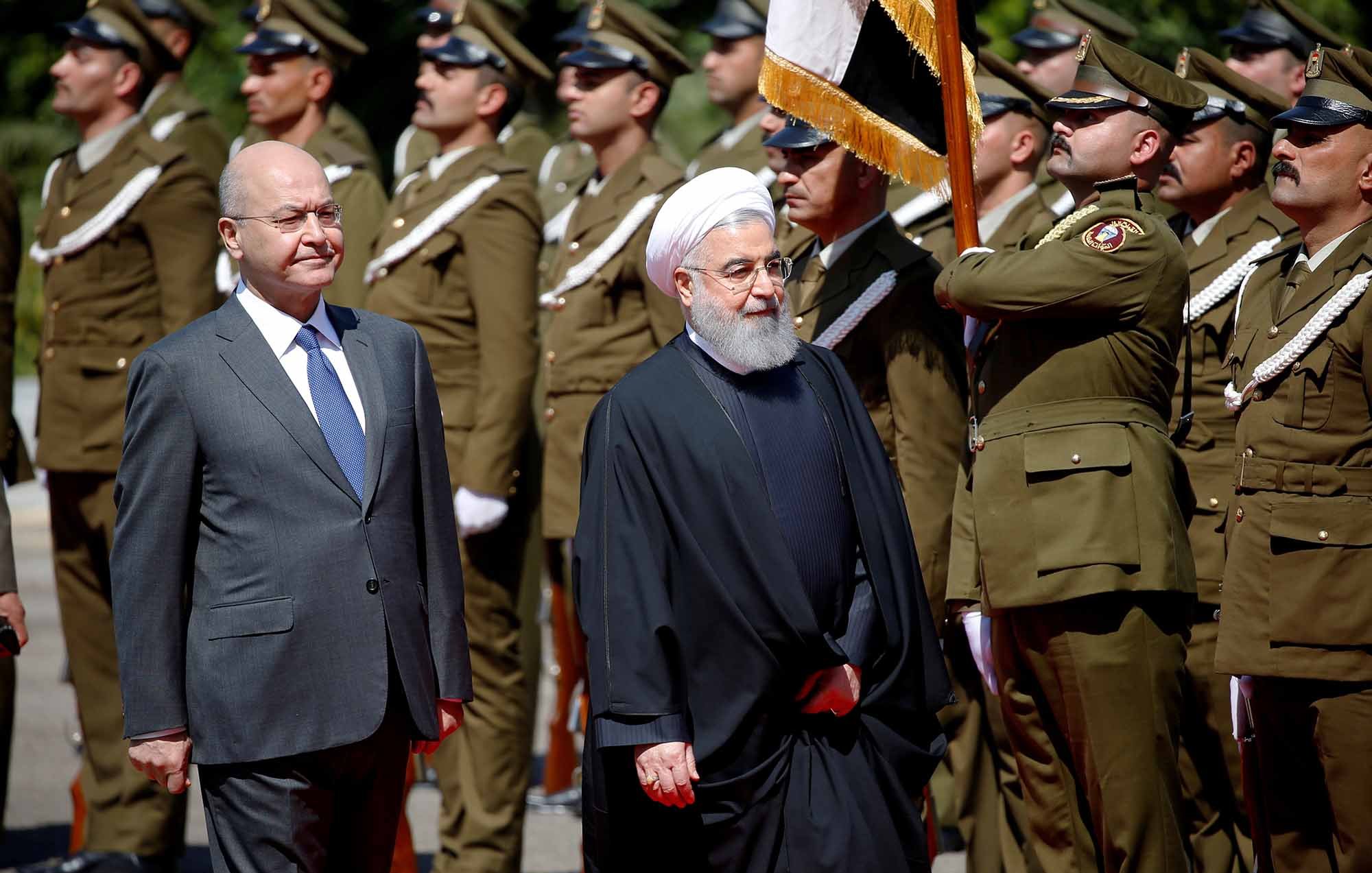 The image shows two men walking in a formal setting, likely during a ceremonial event. One man is dressed in a suit and tie, while the other is wearing a traditional black robe and a white turban. They are surrounded by a group of uniformed soldiers standing at attention, indicating a military or state occasion. The atmosphere appears formal and significant, suggesting a diplomatic meeting or official visit.