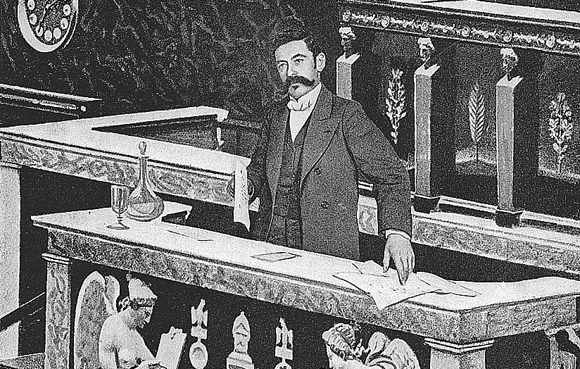 The image depicts a man standing at a podium in what appears to be a legislative or parliamentary setting. He is dressed in a formal suit and has a mustache. The podium is richly decorated, with sculptures of angelic figures and ornate designs. There seem to be documents and a glass on the podium, indicating a formal presentation or discussion taking place. The background suggests an elegant and historical architecture, typical of governmental buildings.