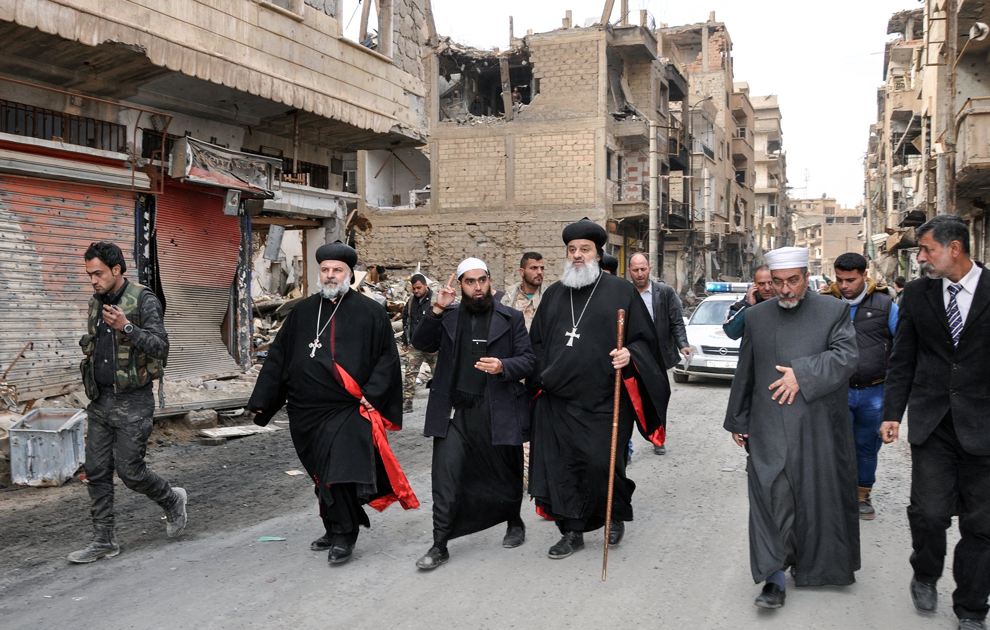 The image shows a group of religious figures walking through a war-torn street. They are dressed in traditional clergy attire, including black robes and crosses, and some are holding staffs. The surroundings are heavily damaged, with buildings showing signs of destruction and debris on the street. In the background, there are a few people and vehicles, indicating a somber atmosphere in a once-bustling area. The overall scene reflects the impact of conflict on the community and highlights the presence of religious leadership amidst the destruction.