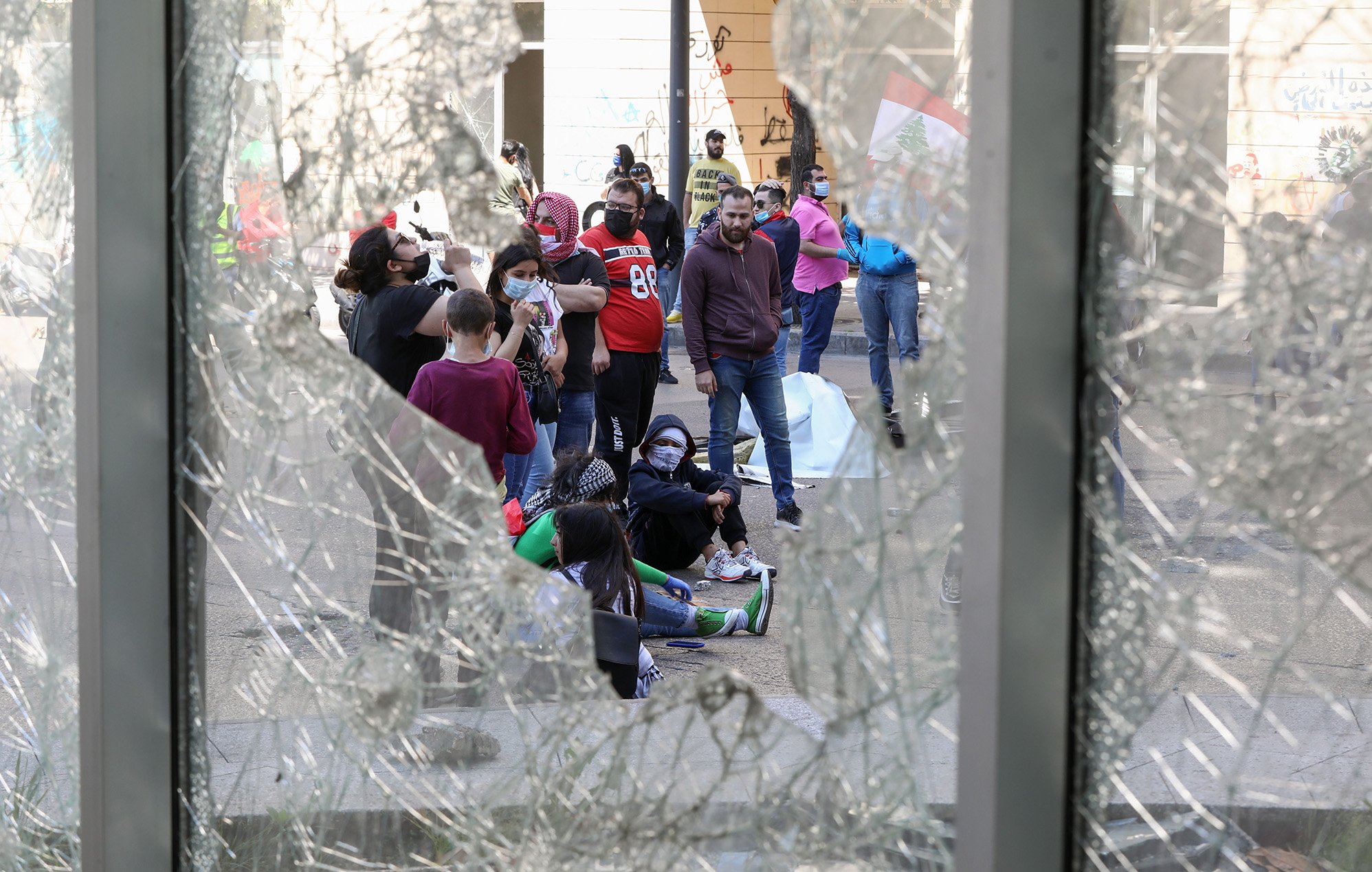 The image shows a group of people gathered on a street, viewed through broken glass. The scene appears to be one of protest or demonstration, as indicated by the presence of individuals wearing masks and the visible graffiti on the walls nearby. Some members of the group are sitting on the ground, suggesting a moment of gathering or reflection amid the chaos. The atmosphere seems tense, reflecting a social or political issue that has drawn this assembly.