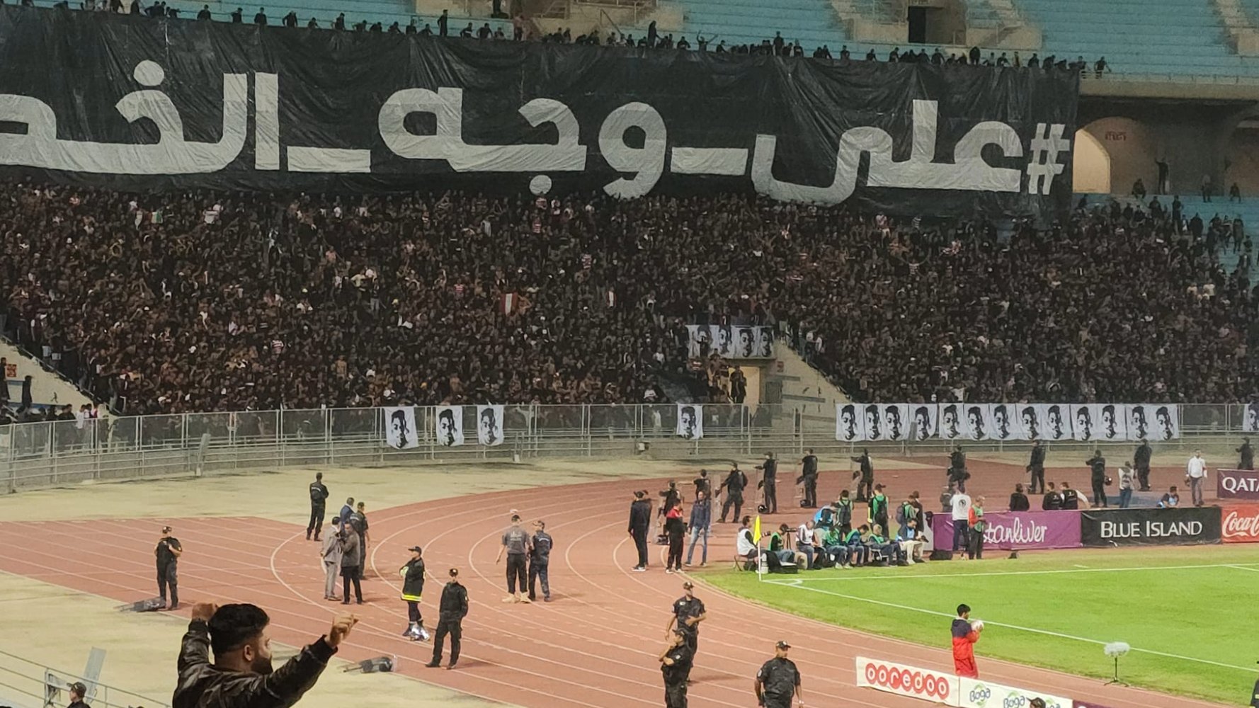A large crowd at a stadium with a black banner and fans cheering passionately.