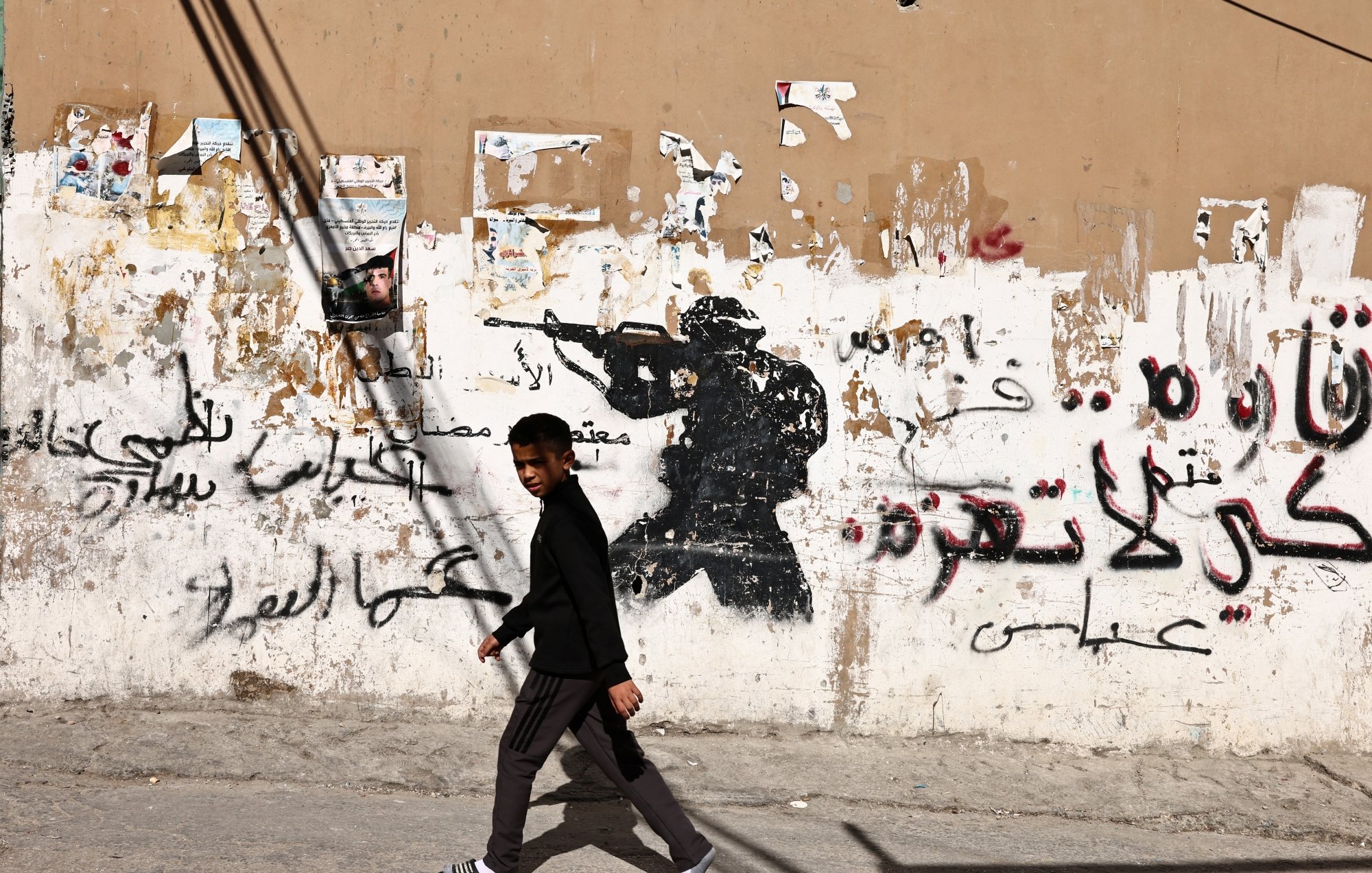 A boy walks past a wall with a soldier graffiti and Arabic text, contrasting street life.