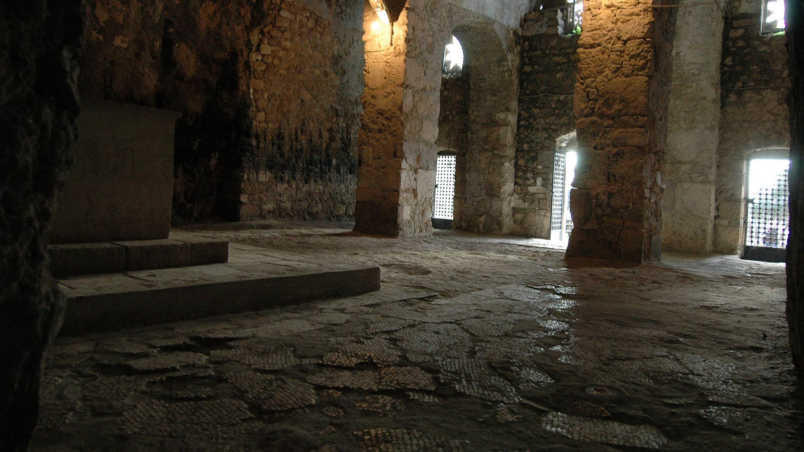 The image depicts an interior space that appears to be a stone chamber or crypt. The walls are made of rough stone, showcasing an ancient architectural style. Light filters in through arched windows, casting a soft glow on the uneven stone floor. There are several columns supporting the ceiling, and a platform or altar can be seen against one wall. The atmosphere is quiet and austere, suggesting a place of historical or spiritual significance.