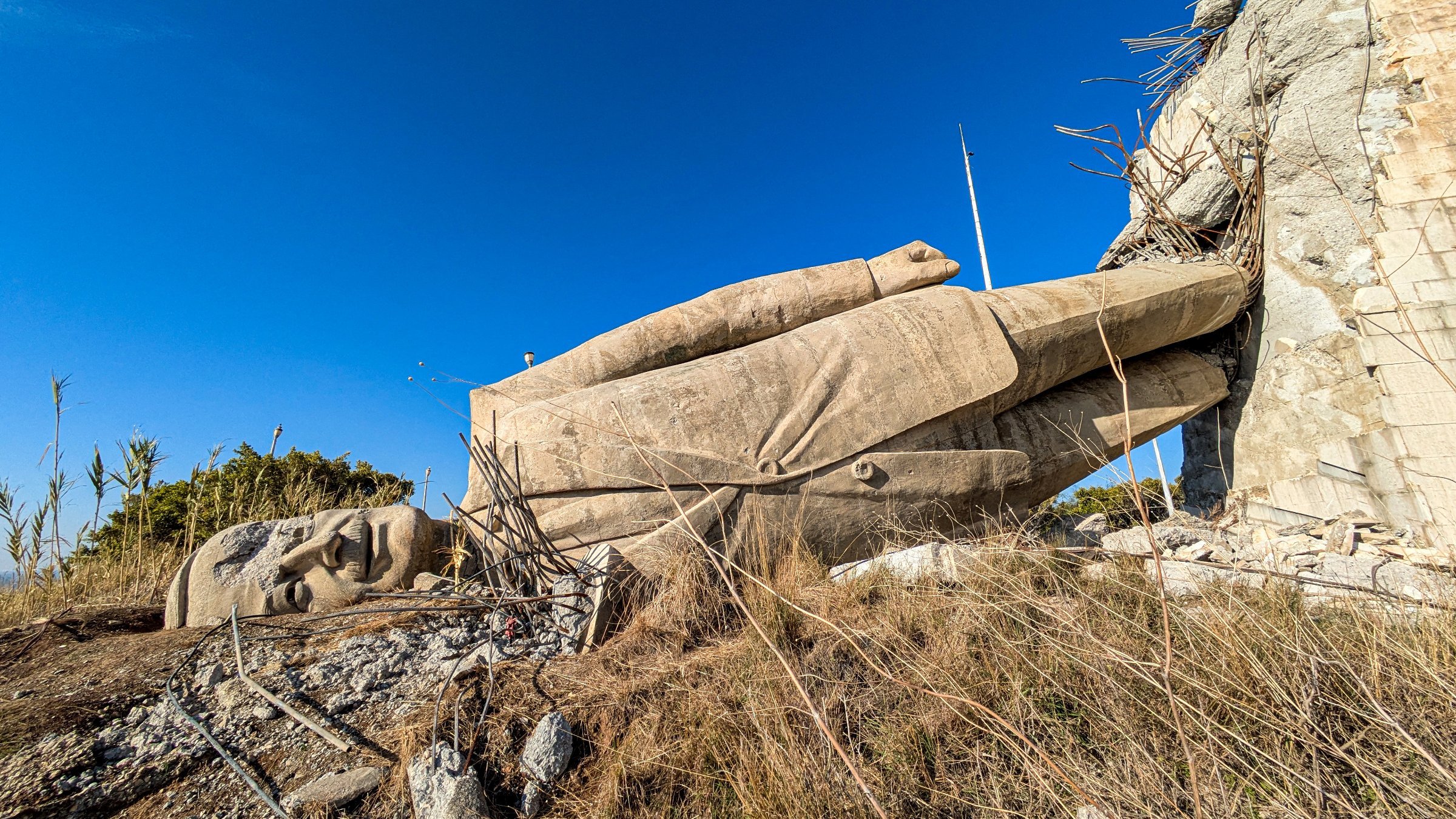 The image shows a large, partially fallen stone statue lying on its side against a rocky background, surrounded by sparse vegetation and a clear blue sky.