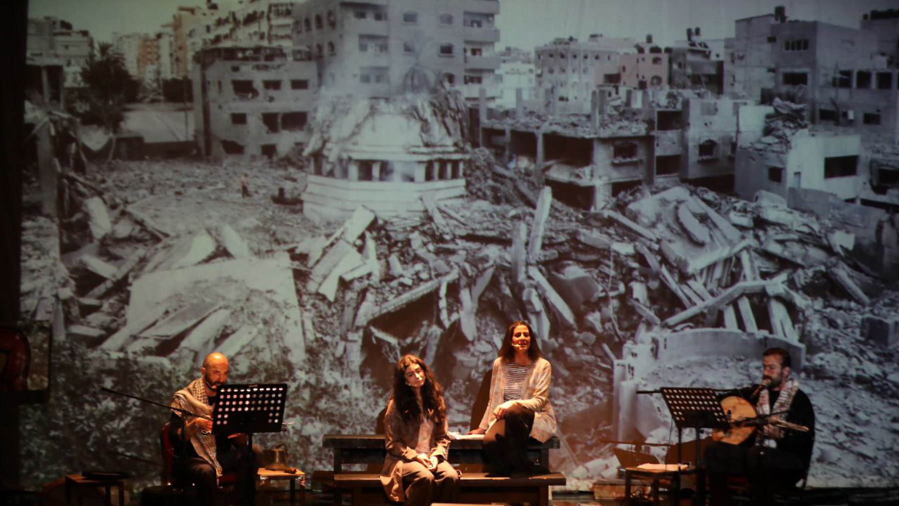 Four performers sit with instruments in front of a large backdrop of war-torn buildings.