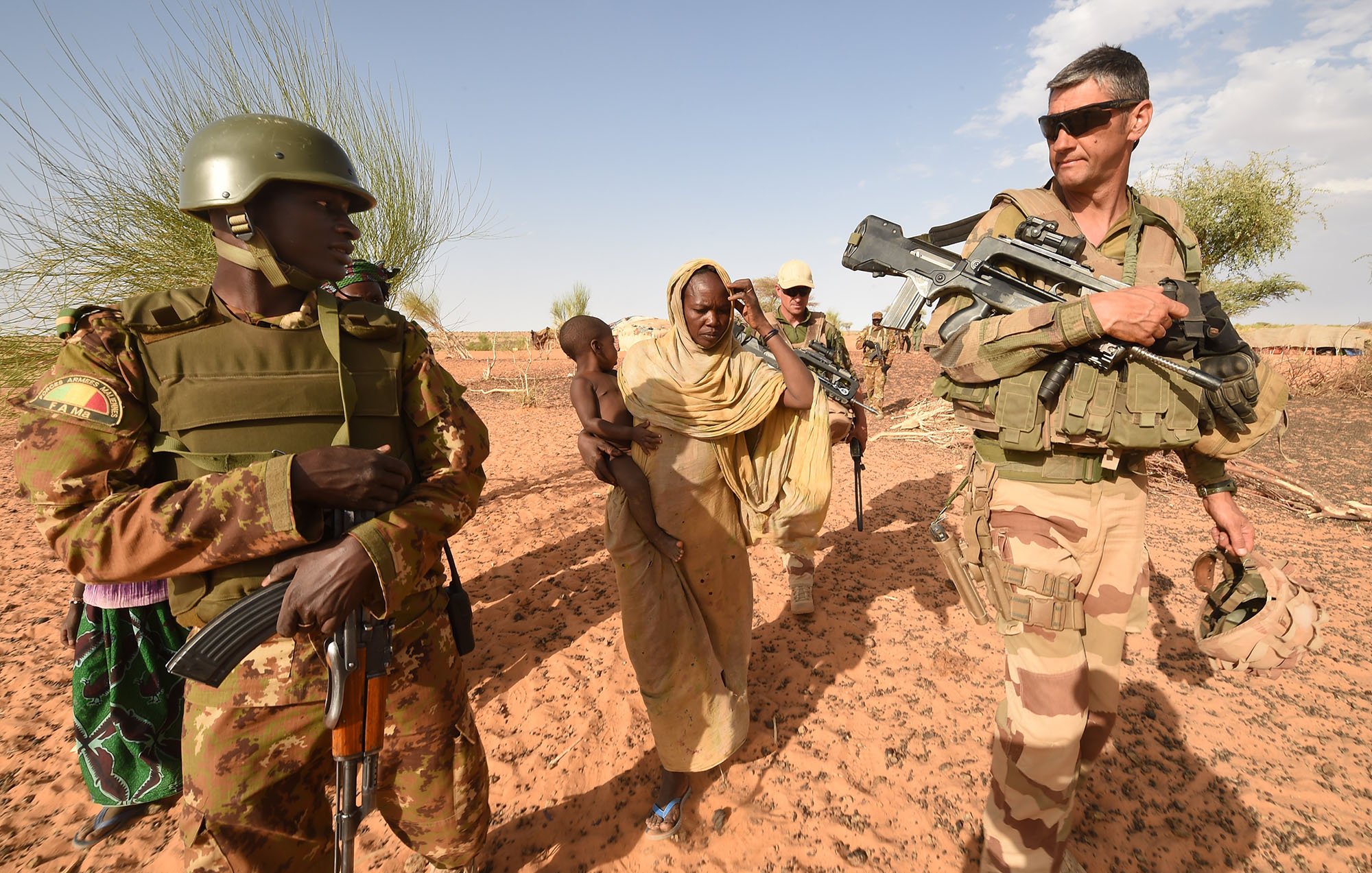 The image shows a scene in a desert setting, featuring military personnel and civilians. On the left, a soldier in a camouflage uniform holds an automatic weapon and wears a helmet, while next to him, a woman in traditional attire carries a young child. To the right, another soldier, dressed in a different military uniform and also armed, is observing the surroundings. The background features sparse vegetation typical of a desert environment. The overall atmosphere suggests a military or peacekeeping operation, possibly in a humanitarian context.