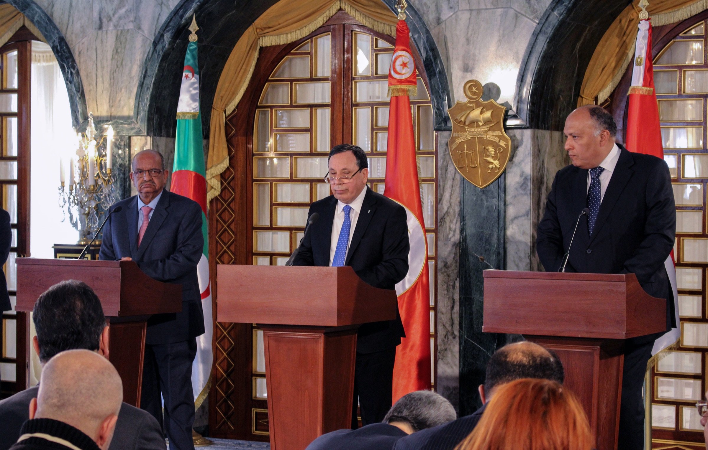 The image depicts a press conference or official meeting involving three individuals standing behind wooden podiums. The man in the center appears to be speaking, while the two flanking him are listening. The backdrop features flags of Algeria and Tunisia, along with a decorative emblem. The setting has an elegant, formal ambiance, likely within a government building. There are several journalists or attendees seated in front, capturing the event.