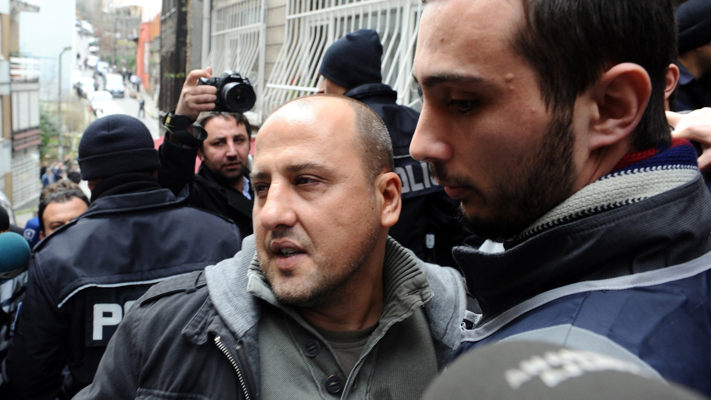 The image depicts a scene with a group of people, likely in a city setting. In the foreground, two men are present. One man, appearing distressed or concerned, is being escorted by another, who seems to be in a position of authority. The background includes police officers and a crowd, indicating a tense situation or an event such as a protest or arrest. The overall atmosphere appears to be one of heightened emotion or urgency.