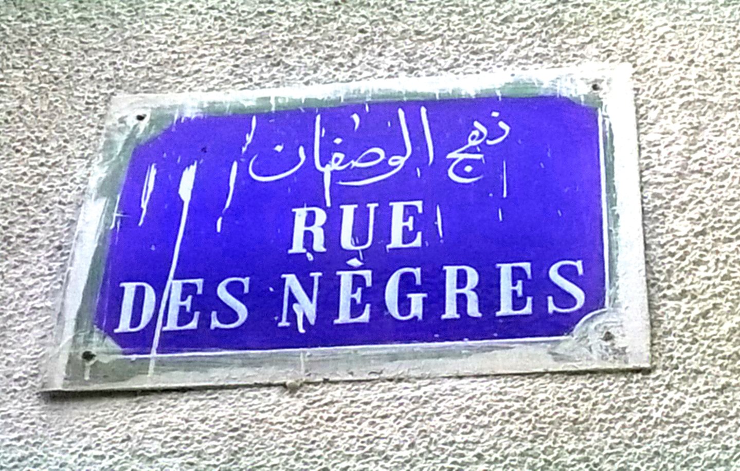The image shows a street sign. The sign is rectangular and features a blue background with white lettering. It includes text in French that reads "RUE DES NÈGRES," along with Arabic script above it. The sign appears to be weathered, with some signs of wear and tear.