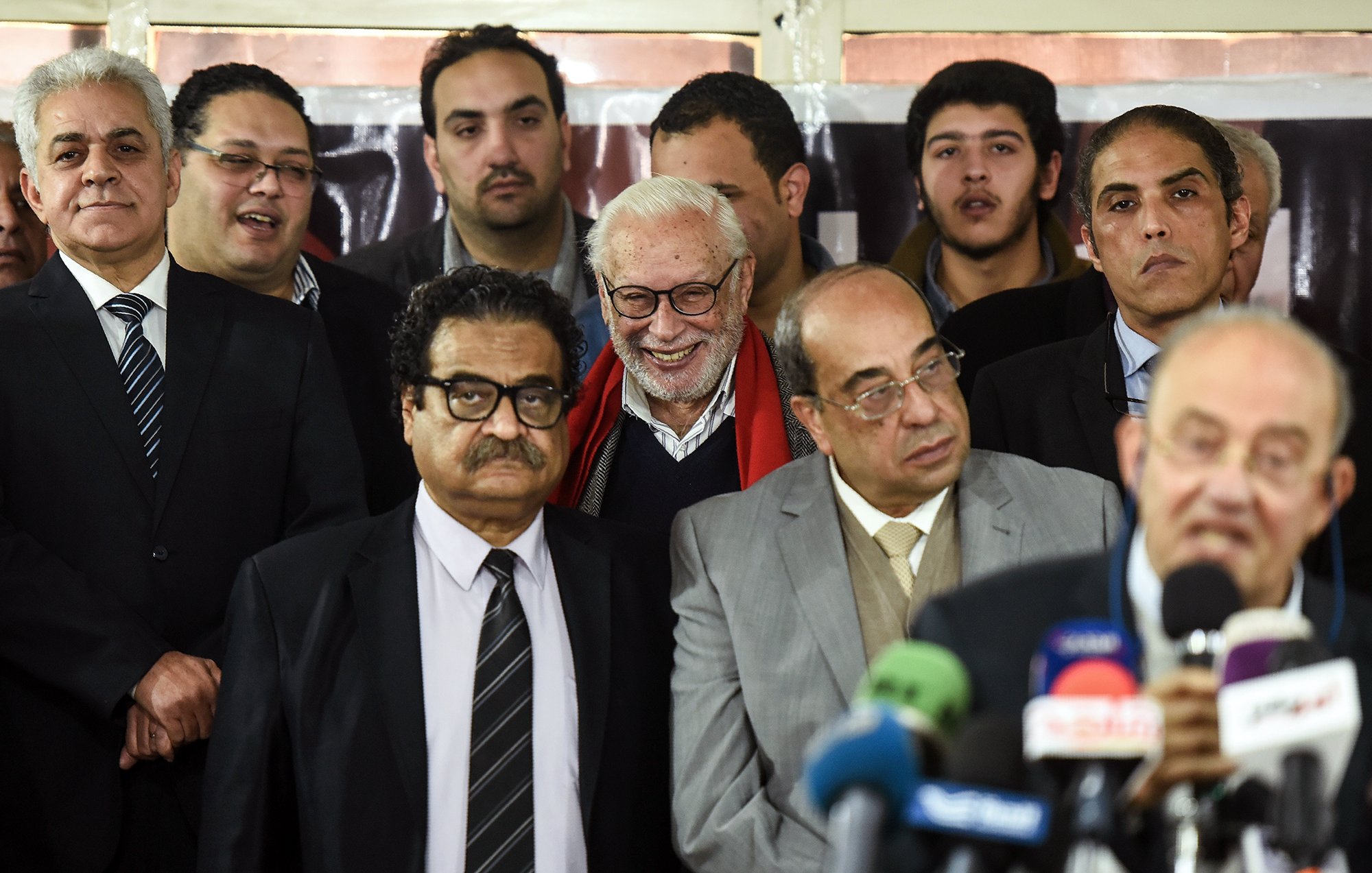 The image features a group of people gathered together, possibly at an event or press conference. There are several men in formal attire, with some wearing suits and ties, while others seem to be casually dressed. In the foreground, one man is speaking into a microphone, while others appear to be listening attentively. There is a mix of expressions among the individuals, including smiles and serious looks. The background includes a banner, suggesting this may be a significant gathering related to a specific topic or announcement.