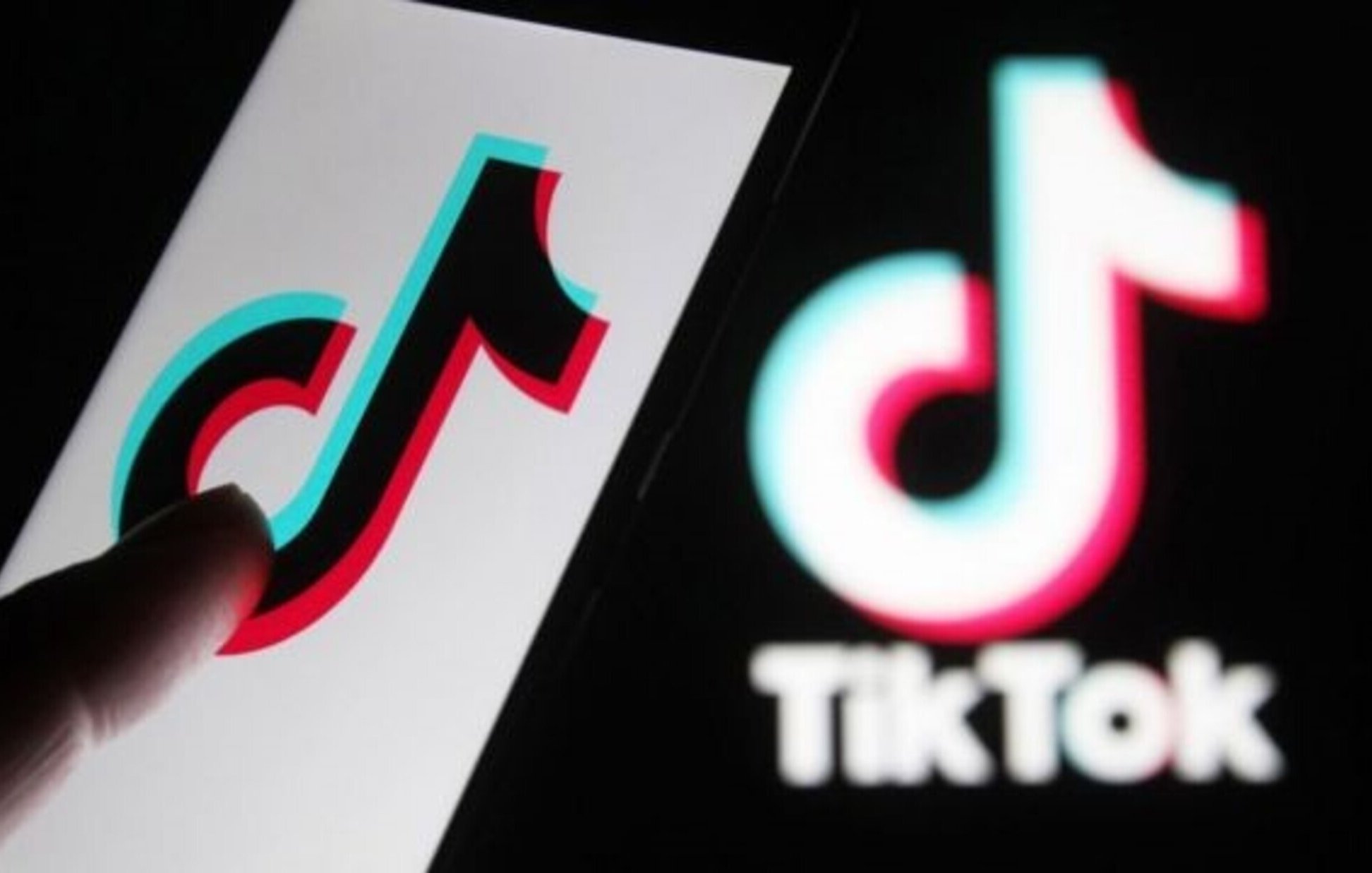 The image features a close-up of a smartphone displaying the TikTok logo, which is designed with vibrant colors—predominantly red, blue, and white. A finger is pointing towards the logo on the screen, while a larger version of the TikTok logo is blurred in the background. The overall focus is on the application's recognizable branding.