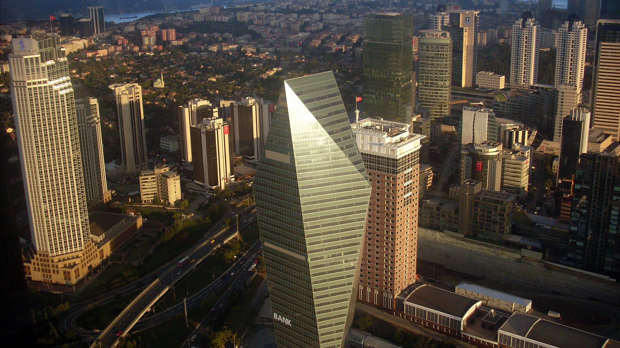 The image depicts a cityscape with a collection of modern skyscrapers and buildings. In the foreground, a distinctive triangular-shaped building stands out, likely serving as a bank or a financial institution. Surrounding it are various other high-rise buildings, indicating a bustling urban environment. The scene appears to be taken from a high vantage point, as it provides a wide view of the city, including green areas and roads. The skyline is clear, suggesting a bright day, with distant mountains or hills visible in the background.