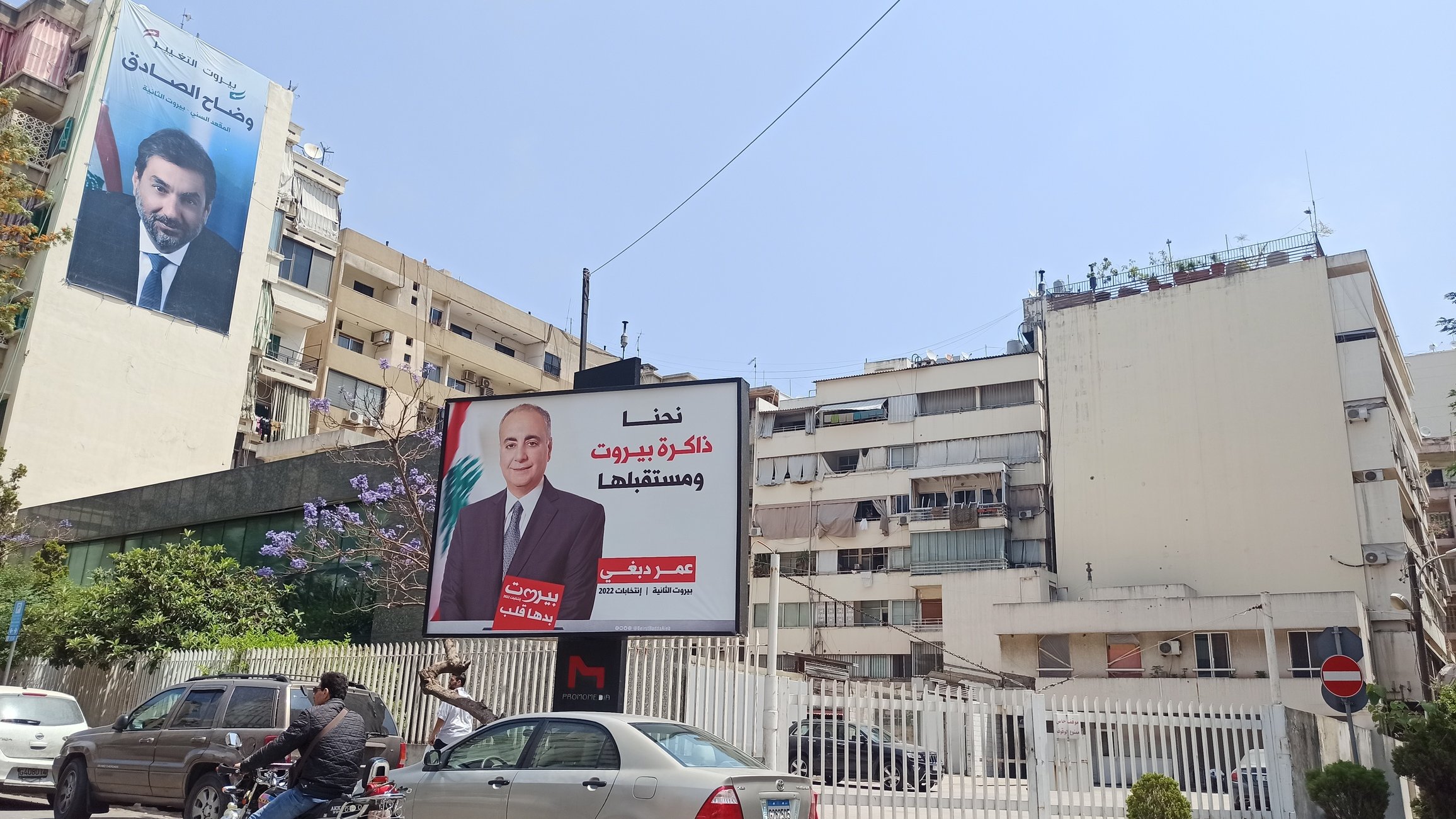 The image shows a street scene featuring a large billboard on a building. The billboard displays a portrait of a man and includes text in Arabic. In the background, there are other buildings, some of which also feature large advertisements. A couple of vehicles, including a motorcycle and a car, are visible on the road. The sky is clear and blue, indicating a sunny day.