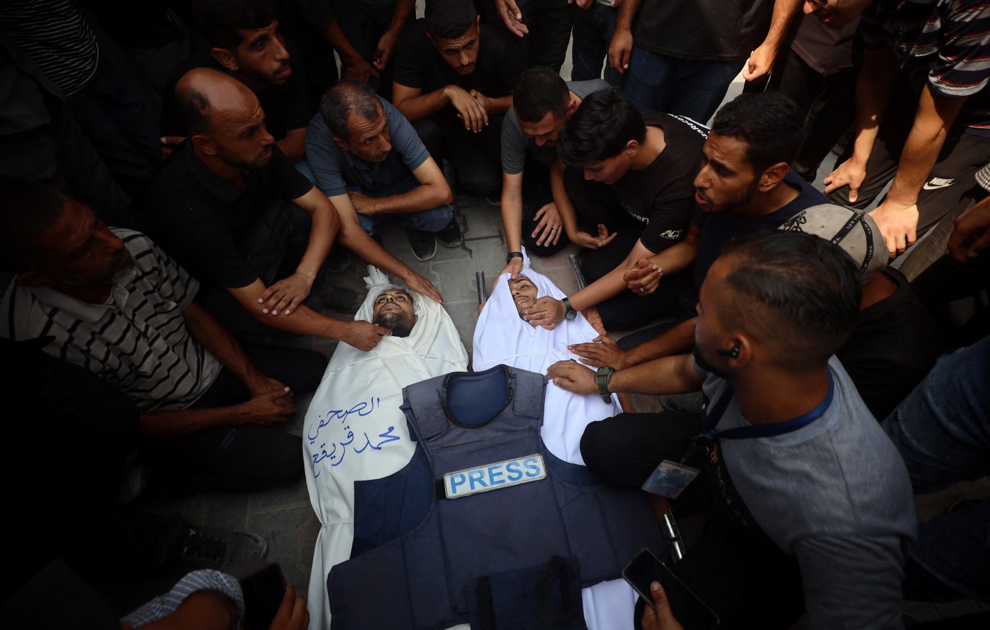 A crowd mourns over a deceased person covered in a white cloth, wearing a press vest.