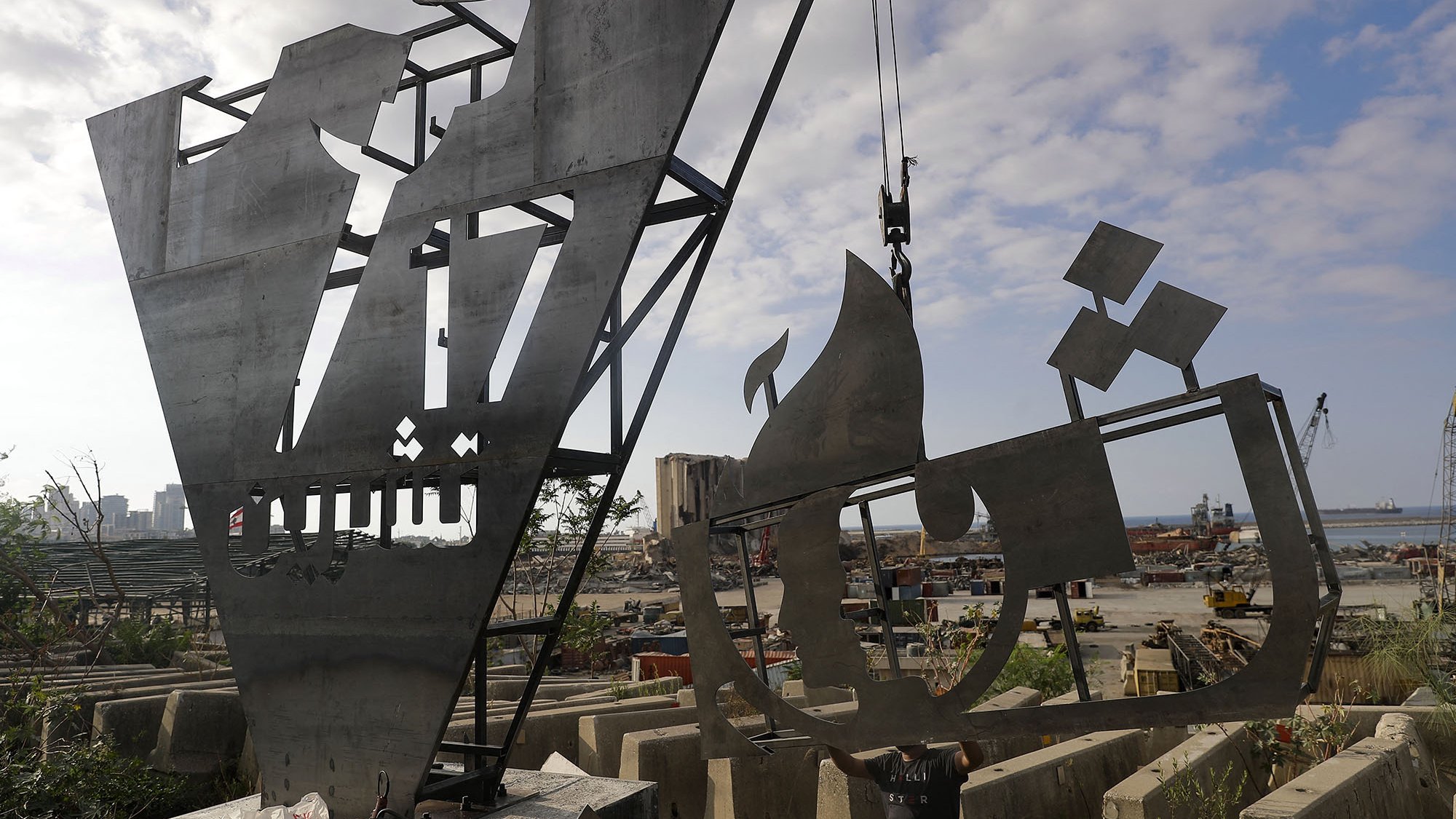 The image depicts two large, metallic sculptures or structures under construction or installation in an industrial outdoor setting. The sculptures show abstract forms, one resembling a stylized letter or shape and the other possibly depicting a ship or vessel. The background features a harbor with cranes and construction materials scattered around, suggesting an active worksite. The sky is partly cloudy, adding to the overall atmosphere of a busy environment.