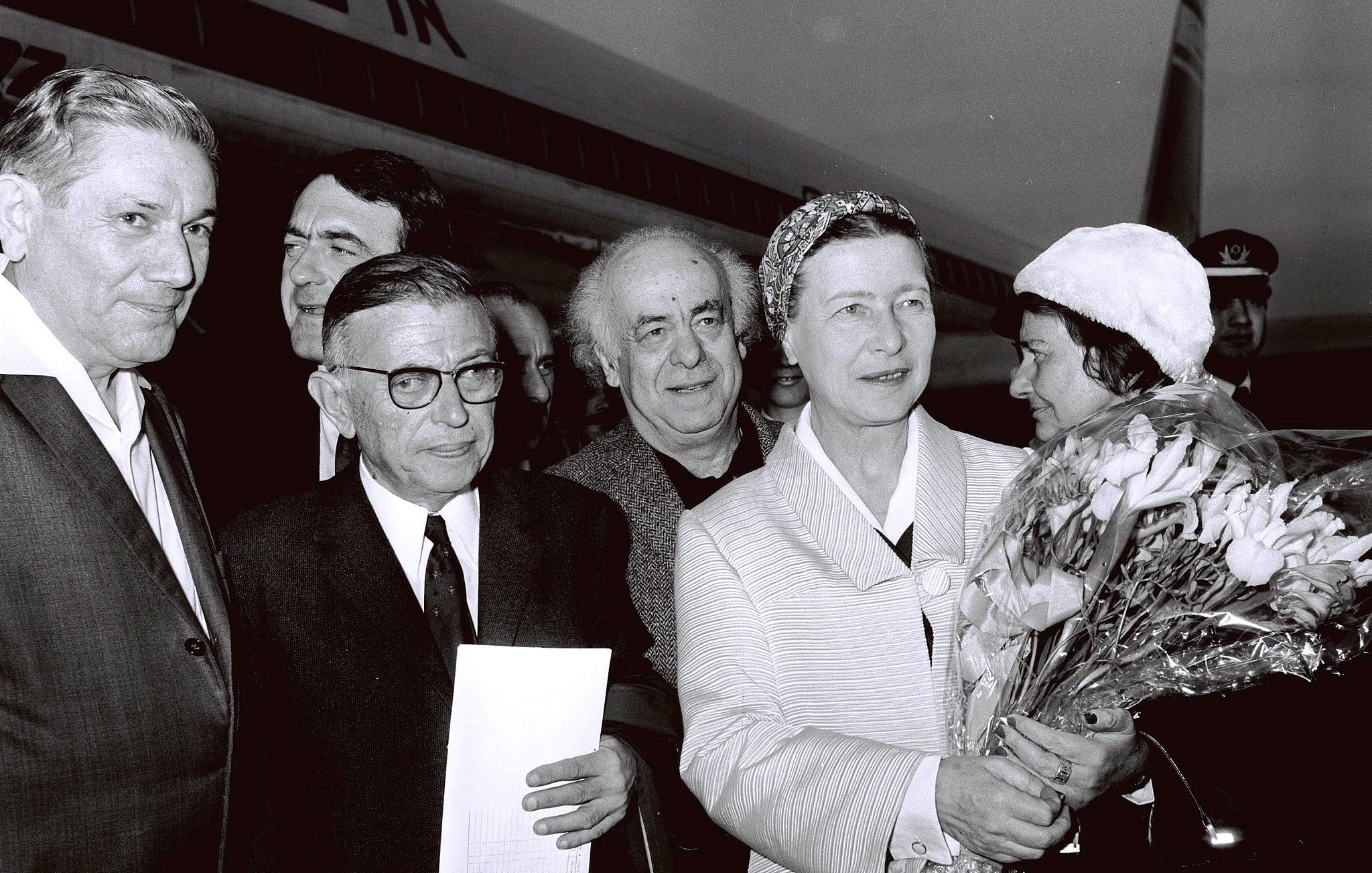 The image captures a group of individuals outside an airplane, likely at an airport. There are six people in the frame, with one woman prominently holding a bouquet of flowers. She appears to be the center of attention, dressed in a light-colored outfit and wearing a headscarf. The men around her are dressed in suits and vary in expressions. The background includes the tail of the aircraft, suggesting a public or ceremonial occasion. The image is in black and white, giving it a classic, historical feel.