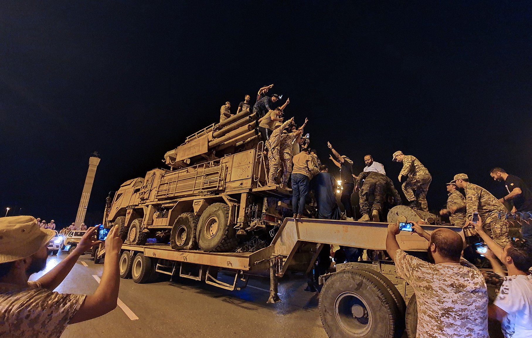 The image shows a large military vehicle, likely a truck, being used in a nighttime setting. A group of people, some in military attire and others in civilian clothing, are gathered around the vehicle. Some individuals are climbing on top of it, while others are taking photos with their phones. The atmosphere appears lively, and there is a sense of celebration or excitement among the crowd. The scene is illuminated by artificial lights, contributing to the nighttime ambiance.