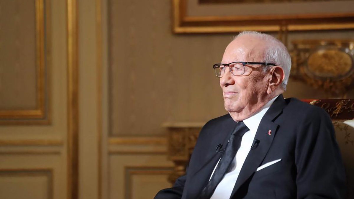 The image depicts an elderly man sitting in a formal setting. He is wearing glasses and a black suit, along with a black tie. The background features tasteful decor with intricate designs and warm colors, suggesting an elegant environment. The man appears to be engaged in thought or reflecting, with a calm and serene expression on his face.