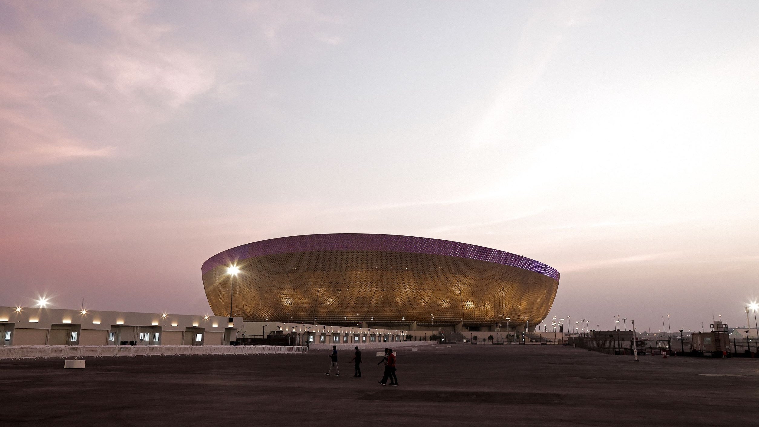 The image shows a large, modern stadium with a unique, oval design. The stadium features an exterior of golden-hued materials, giving it a striking appearance, especially during twilight. There are a few people walking in the foreground, and the sky is painted in soft pastel colors, suggesting either dawn or dusk. The stadium is well-lit, with lights illuminating its structure and the surrounding area. The overall atmosphere is calm and serene, with a sense of anticipation for an event.