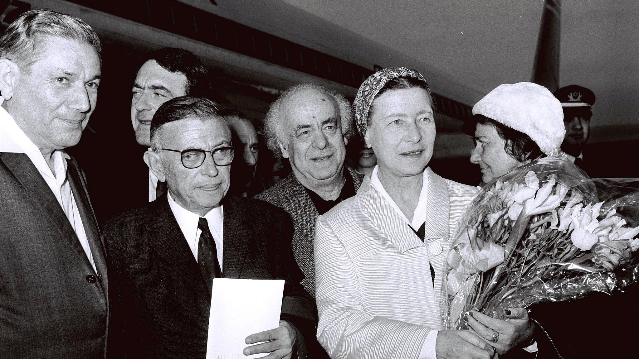The image captures a group of individuals outside an airplane, likely at an airport. There are six people in the frame, with one woman prominently holding a bouquet of flowers. She appears to be the center of attention, dressed in a light-colored outfit and wearing a headscarf. The men around her are dressed in suits and vary in expressions. The background includes the tail of the aircraft, suggesting a public or ceremonial occasion. The image is in black and white, giving it a classic, historical feel.