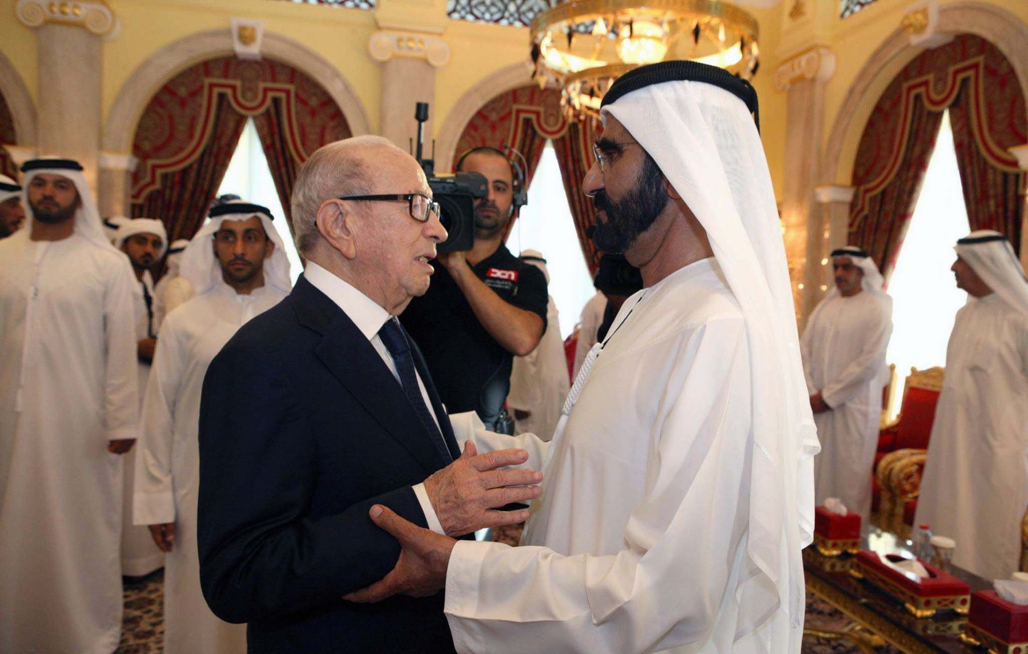 The image depicts two men engaging in conversation, likely in a formal setting. One man is dressed in a dark suit and glasses, while the other is in traditional Emirati attire, including a white kandura and headscarf. They appear to be having a cordial exchange, with the background showing a lavishly decorated room, including red curtains, ornate furniture, and a gathering of individuals in similar traditional attire. The atmosphere seems formal and respectful.