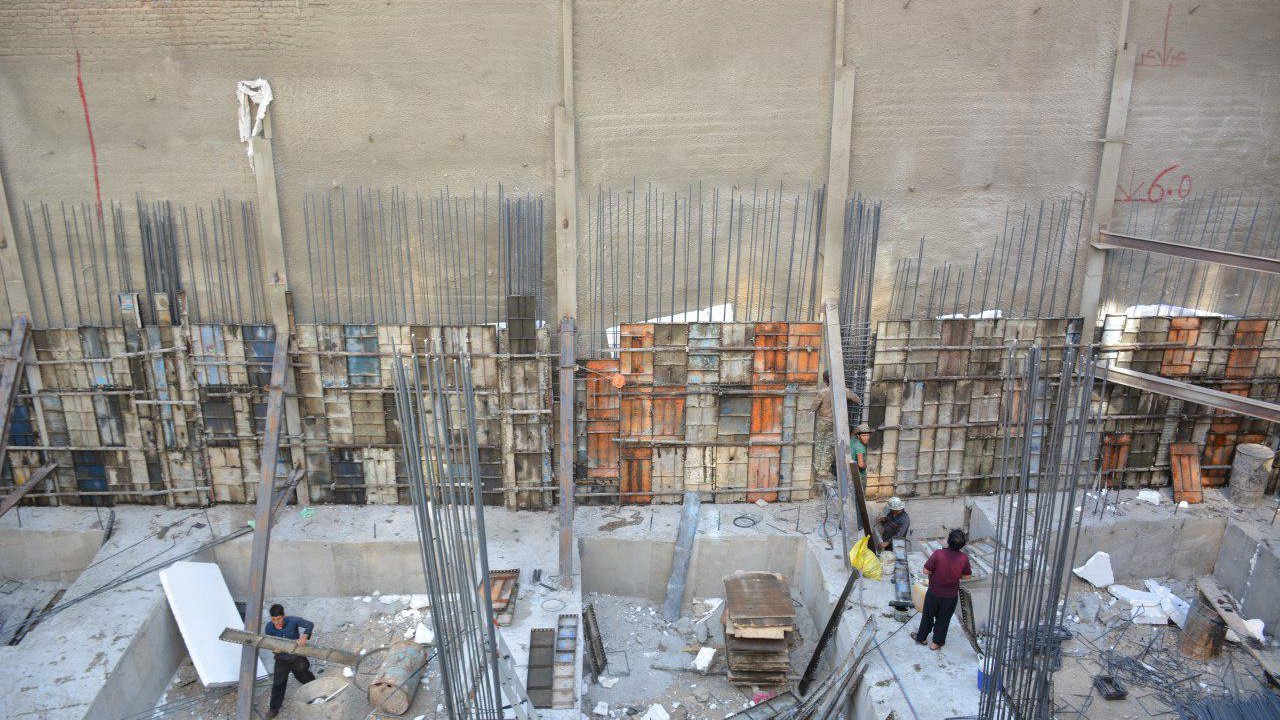 The image shows a construction site with several workers engaged in various activities. There are exposed concrete walls, rebar (steel reinforcement bars) standing upright, and wooden forms for pouring concrete. The area appears to be a deep excavation where preparations for building are underway. Some materials like wood and steel are scattered around, while a few workers are busy, possibly organizing or mixing materials. The overall scene reflects typical construction activities in an urban setting.