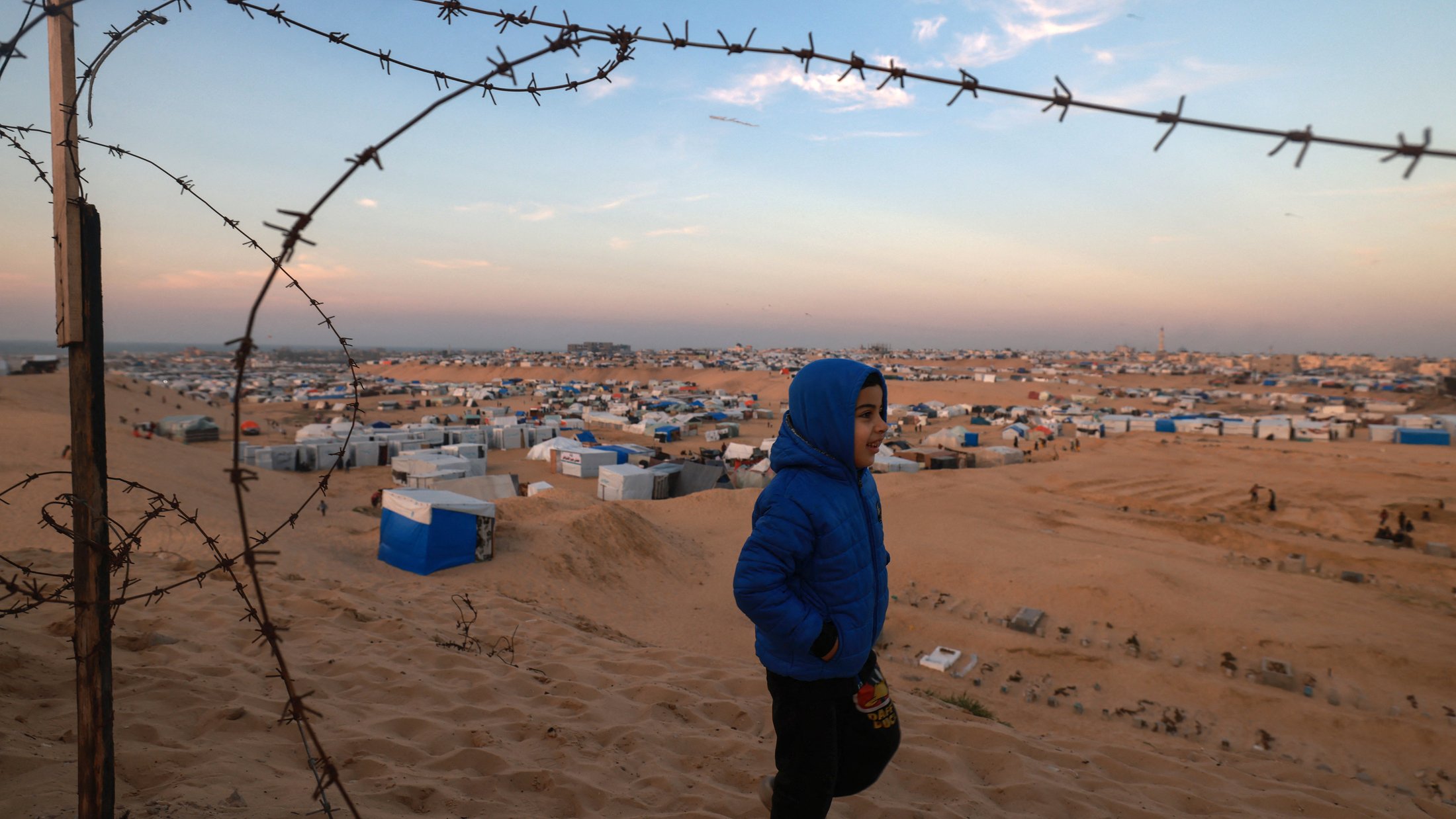The image shows a child wearing a blue jacket walking along a sandy landscape. In the background, there are numerous temporary shelters, likely tents or makeshift homes, scattered across the terrain. The sky is a mix of soft colors, indicating either dawn or dusk. A fence topped with barbed wire runs along the foreground, adding a sense of restriction or boundary to the scene. The overall atmosphere conveys a sense of hardship, amidst the child's movement.